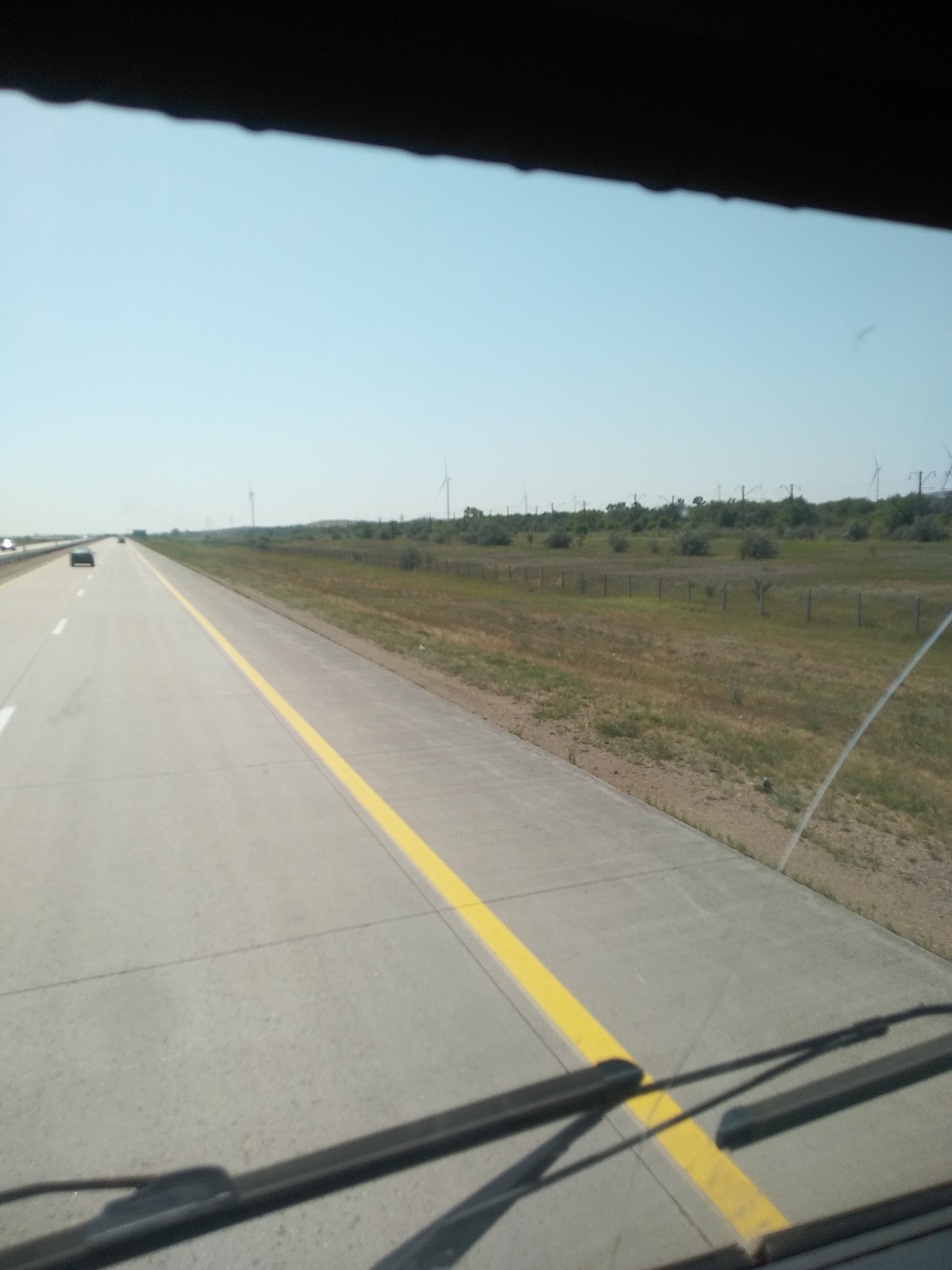A straight, multi-lane highway stretches into the distance under a clear, light blue sky. The road surface is concrete with a bright yellow line dividing lanes. Several cars are visible on the highway, some in the distance. To the right of the highway, a grassy field with scattered bushes and trees transitions to rolling hills. Several wind turbines are visible on the horizon. The foreground shows the windshield wipers of a vehicle, casting shadows on the road. The scene appears to be a rural highway, likely outside a populated area.
