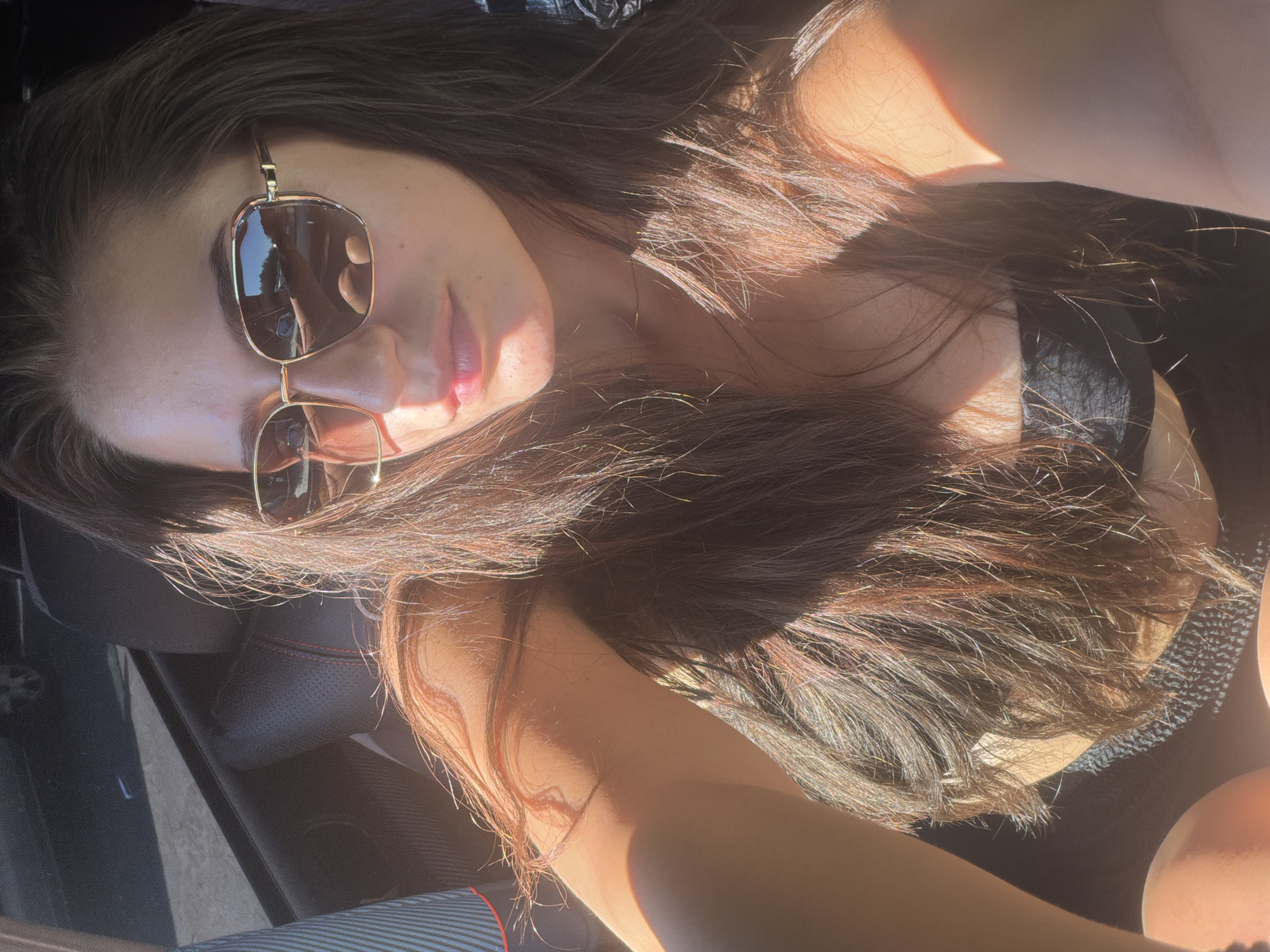 The image shows a young woman with long, dark brown hair and sunglasses sitting in the passenger seat of a car. The sun is shining brightly, casting strong shadows and highlights on her face and hair. She is wearing a black strapless top. The interior of the car is visible, including the black leather seats with red stitching. The background is blurred, but appears to be an outdoor urban setting, possibly a street in Burgas, Bulgaria.