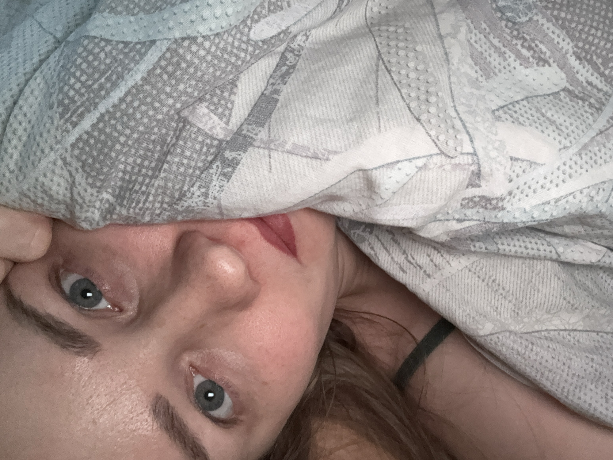 An intimate, close-up shot captures a person partially hidden beneath a light gray and white patterned blanket, oriented diagonally across the frame. The subject, appearing to be a woman, has fair skin, distinct blue-grey eyes looking directly at the viewer, and arched eyebrows. Her nose is visible, and her lips are painted with a soft pink or red lipstick. A strand of her reddish-brown hair is visible to the right of her face, and a dark strap, possibly from a piece of clothing, peeks out near her shoulder.
The blanket, which covers most of her head and body, features an abstract design with areas of small dots and linear patterns. The lighting is soft and even, suggesting an indoor setting, likely a bedroom. The overall impression is one of a quiet, private moment, perhaps waking up or relaxing in bed in Chaikovsky, Russia. There are no strong emotional cues, rather a neutral or slightly curious expression. No text is visible in the image.