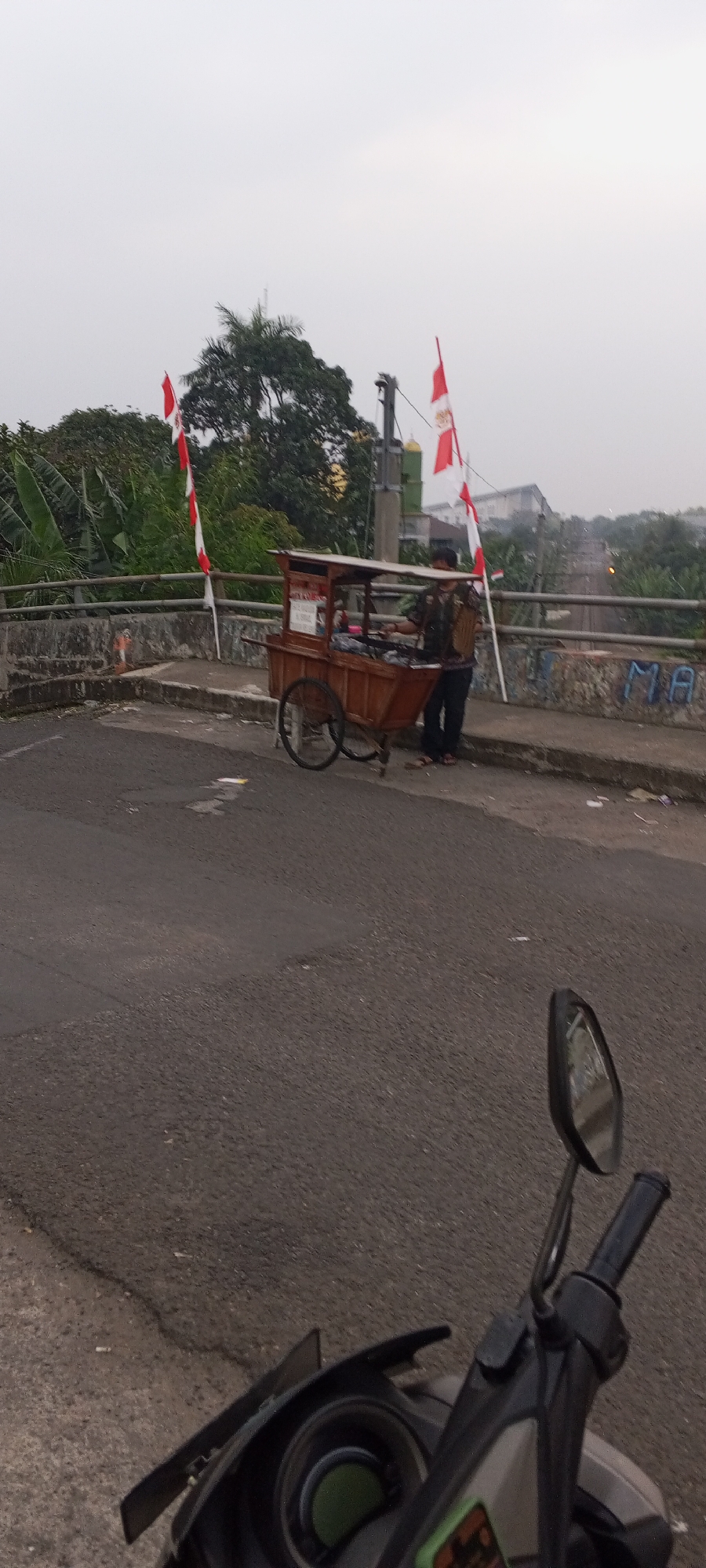The image depicts a street scene in Cibinong, Indonesia, featuring a street vendor's cart on the side of the road. The cart is made of wood and has a covered top, with two people standing near it. One person, wearing a patterned shirt and dark pants, appears to be interacting with the cart. The other person, in a camouflage-patterned vest and dark pants, stands beside them. Several Indonesian flags, red and white, are displayed on poles around the cart, suggesting a national celebration or event. In the foreground, the handlebars and mirror of a motorcycle are visible, indicating the perspective is from a rider. The background shows lush greenery, including banana trees, and a bridge structure. The sky is overcast, and the road surface shows signs of wear and tear with some debris scattered around.