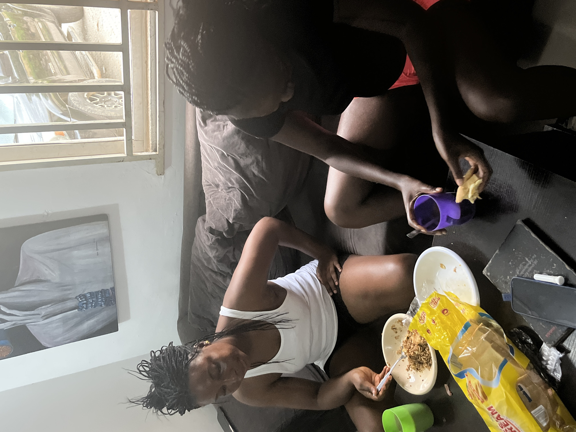 The image shows two young Black women seated on a couch in an indoor setting, likely a living room in Abuja, Nigeria. One woman is wearing a black tank top and the other a white tank top. They are positioned around a dark coffee table cluttered with food bowls, a plastic cup, and a yellow bread bag. One woman is actively eating from a bowl with a fork, while the other holds a purple cup and appears to be reaching for something on the table. The overall atmosphere seems casual and relaxed, possibly indicating a mealtime or a break.
Visible text includes "CLASSIC CREAM" on the yellow bread bag. A painting of a person in light-colored clothing is on the wall to the left. The scene is lit by natural light coming from a window with horizontal blinds, suggesting it is daytime. Outside the window, car tires and parts are visible, hinting at a car or a repair area outside the building. The image is oriented vertically.