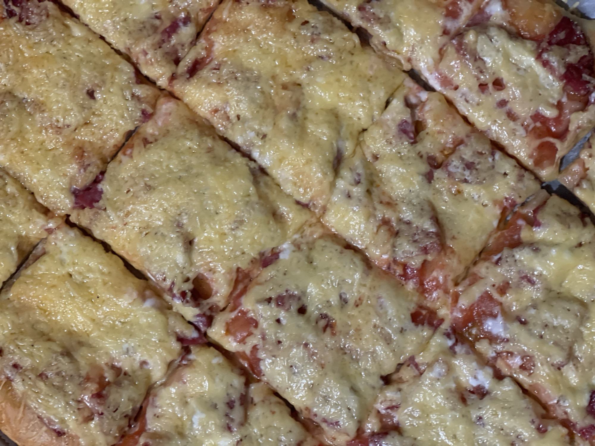 A close-up, overhead shot shows a square-cut pizza with melted cheese. The pizza appears to be topped with some kind of red ingredient, possibly tomato or pepperoni. The lighting is warm, creating a slightly glossy sheen on the cheese. The image is tightly cropped, focusing solely on the texture and appearance of the pizza. The location is listed as Unknown, Unknown.