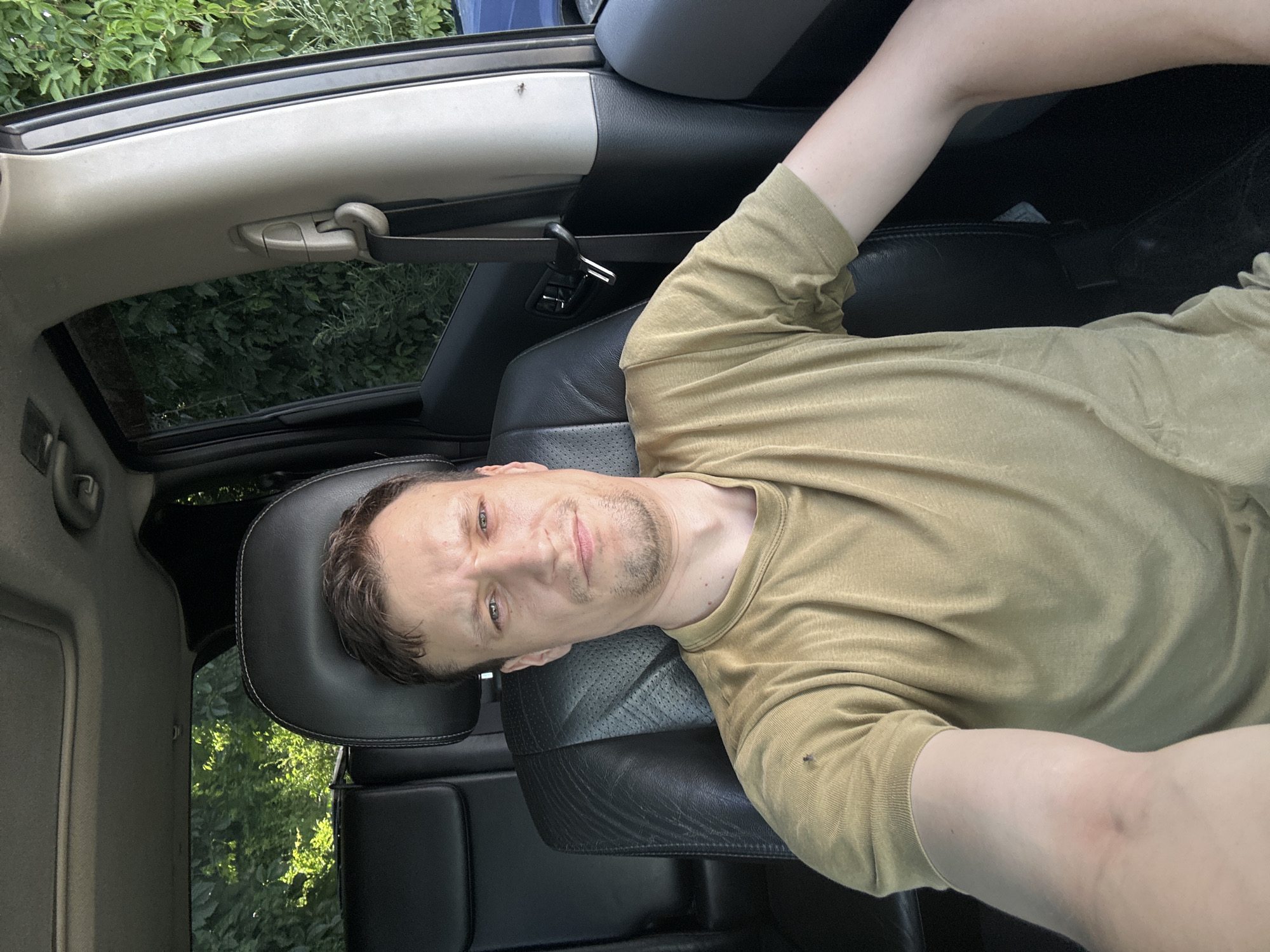 A Caucasian male is sitting in the driver's seat of a dark-colored SUV. He is wearing a light brown t-shirt and has short, dark brown hair and a short beard. The car's interior is visible, including leather seats and a sunroof. Outside the car, lush green foliage is visible, suggesting a natural or park-like setting within or on the outskirts of Shatava, Ukraine. The lighting indicates it is daytime, likely late morning or early afternoon.