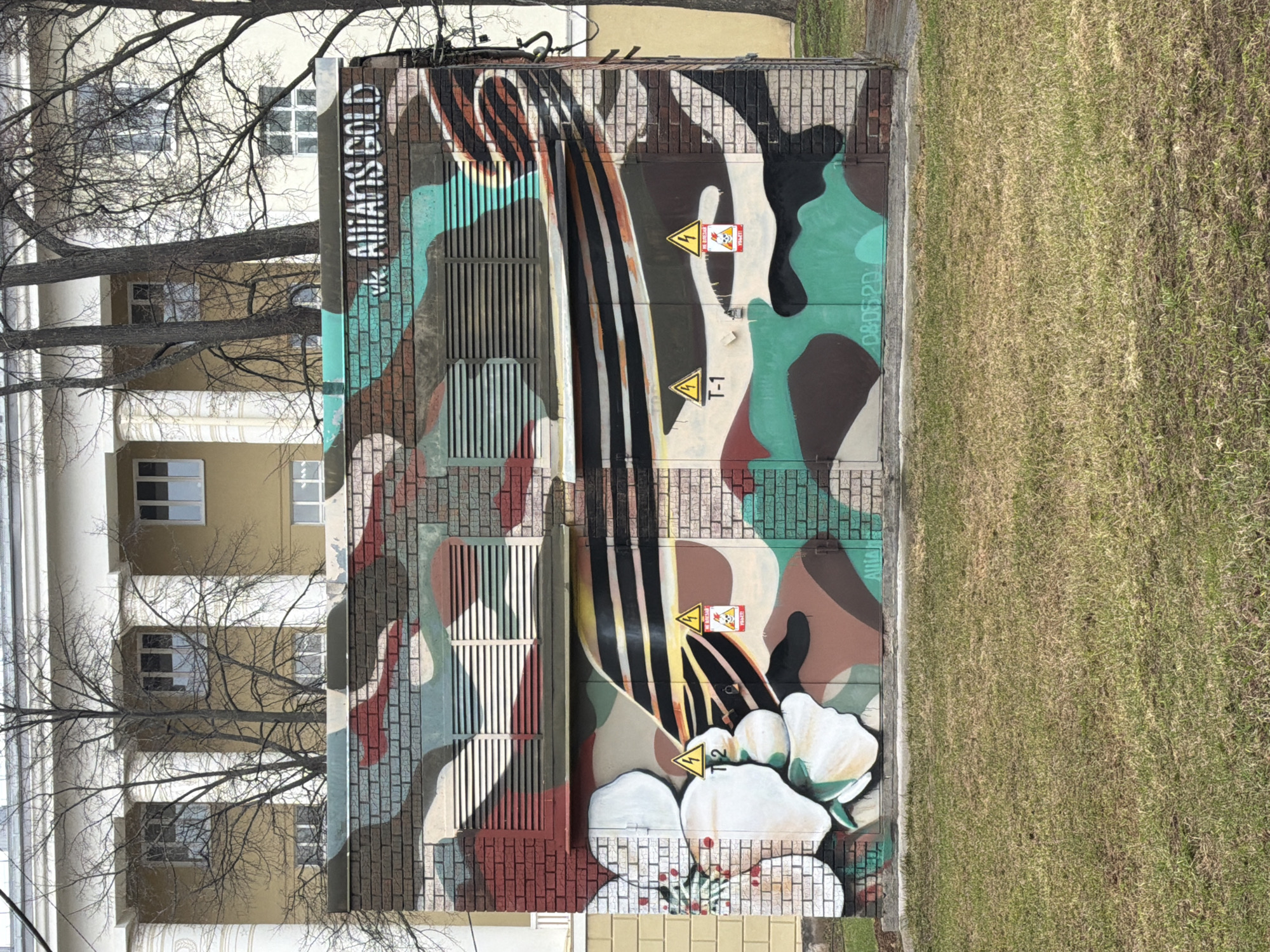 An electrical utility box or small building in Votkinsk, Russia, is the central subject, entirely covered in a vibrant graffiti mural. The artwork features a camouflage-like pattern in various shades of brown, green, black, white, light blue/teal, and red, with some areas designed to resemble brickwork. A prominent abstract element, possibly depicting a stylized violin or cello bow with strings, or a flowing ribbon, sweeps across the center in black and white stripes. Below this, white, cloud-like or floral shapes are painted near the base.
Several caution signs are incorporated into the mural: yellow triangles with black lightning bolts, labeled "T-1" and "T-2," indicating electrical hazards. One sign also includes a red square with a skull and crossbones and the word "ОПАСНО!" (DANGER!).
Visible text includes "VK AllMANS GOLD" painted vertically on the left side of the box, and a smaller tag "08D520" on the right.
To the left of the utility box, a large, bare deciduous tree stands, its intricate branches silhouetted against a multi-story building in the background. This building, likely an apartment or office block, features a light-colored facade with regularly spaced windows and some classical architectural elements like columns. To the right of the utility box is a patch of sparse, dry-looking grass. The scene appears to be captured during the daytime under clear weather conditions, with no specific human activity taking place.