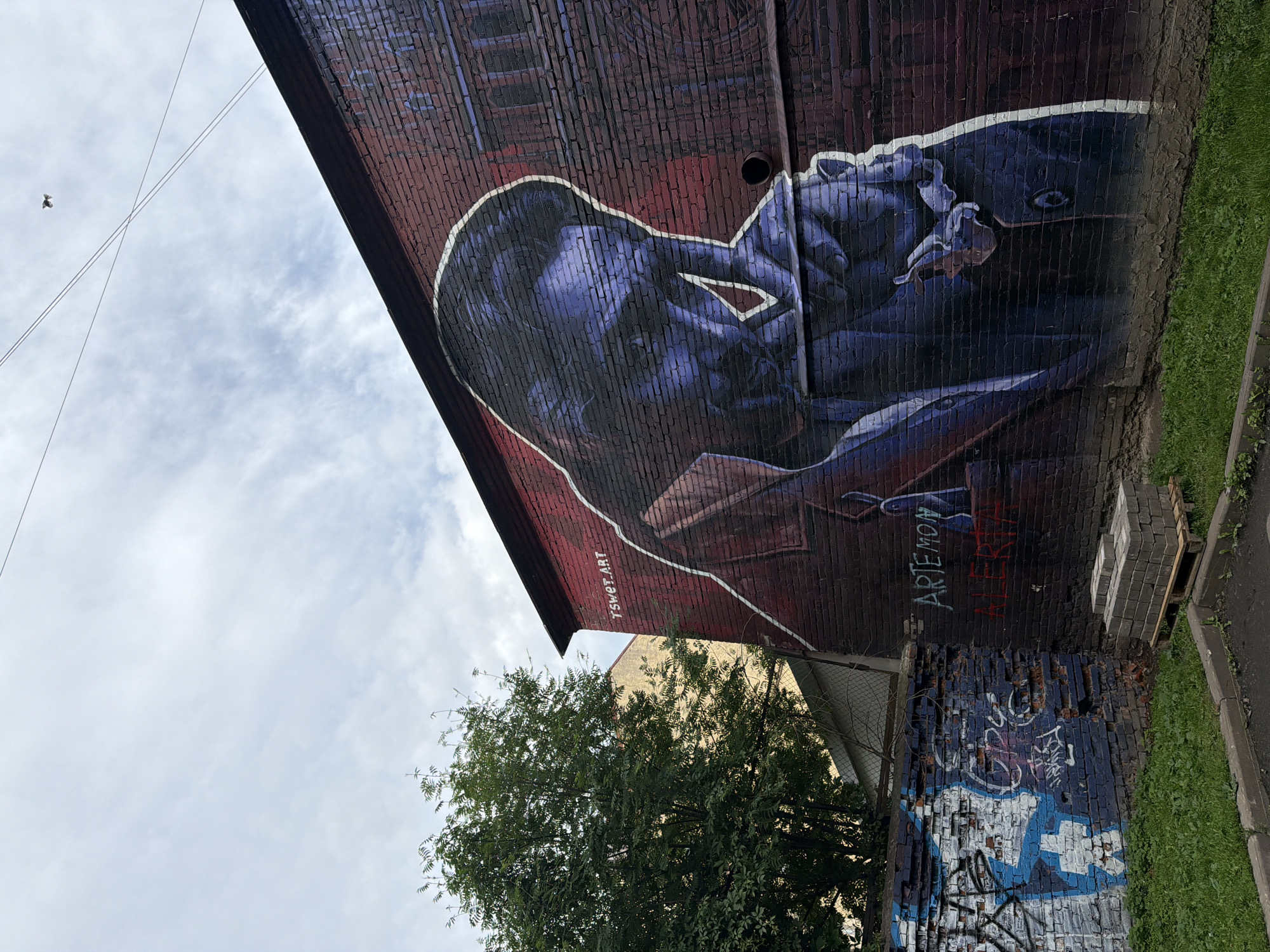 In the city of Unknown, Unknown, a street mural adorns the side of a brick building. The mural depicts a close-up of a person's face and upper body, rendered in shades of blue and purple, with a thin white outline highlighting the form. The person is depicted with their hand near their mouth, in a pensive or contemplative pose. Above the mural, the text "TSWET.ART" is visible in white lettering. To the left of the main mural, a section of a different brick wall is covered in graffiti, featuring blue and white spray paint. In the foreground, a patch of green grass borders a paved area, and a small pile of grey bricks rests on a wooden pallet. A large green tree stands to the left of the building, its branches reaching towards the sky. The sky above is cloudy, with power lines crossing the frame. A small, dark object, possibly a bird or insect, is visible against the sky in the upper right quadrant.