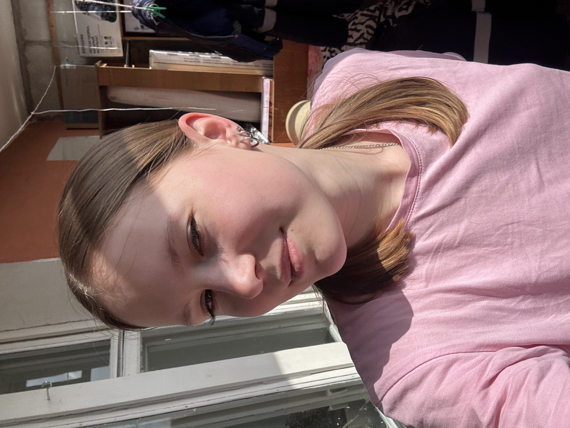 A young woman with light brown hair is indoors, wearing a pink long-sleeved shirt and a silver necklace. She has multiple earrings in her left ear. She is positioned near a window with white frames. Outside the window is a faded orange wall or building. Behind her and to the left are shelves, boxes, and hanging clothes. The image may have been taken in Chaikovsky, Russia, given the request. The woman appears to be looking at the camera. The lighting suggests it's daytime.