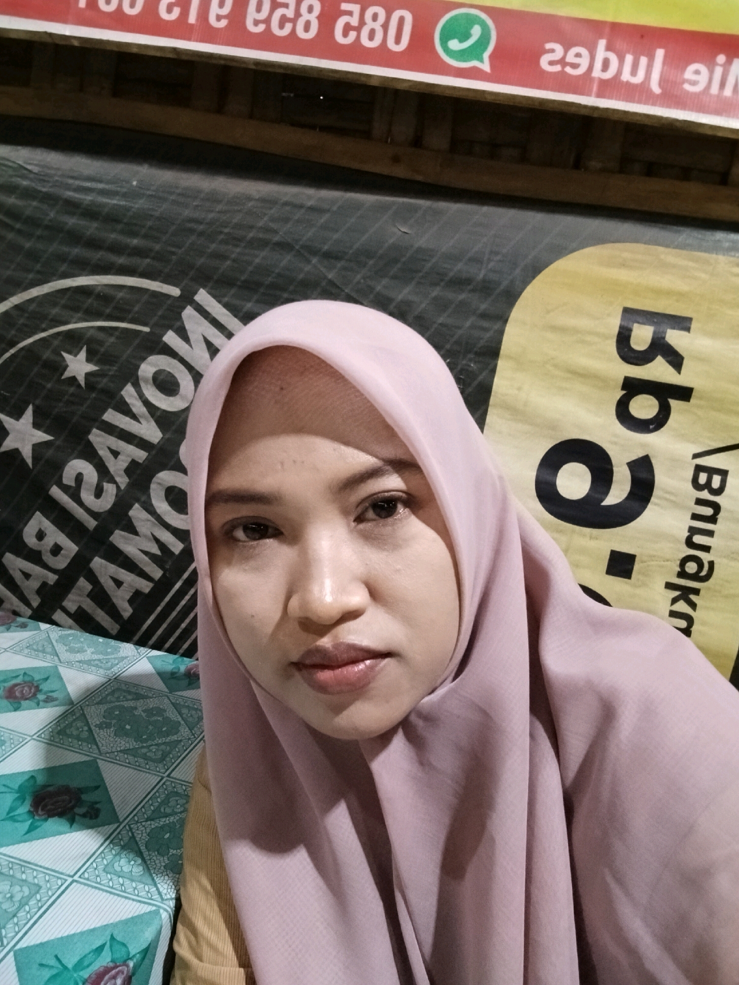 A headshot shows a woman wearing a light pink hijab. She is looking towards the camera with her eyes open and her lips slightly parted. The background appears to be outdoors, with a dark green tarp visible behind her head. To the left of the woman, a portion of a dark green banner with white text is visible. The text includes what appears to be "INOVASI BA TAMO". To the right of the woman, a yellow banner with large black text is partially visible, featuring what looks like "bb 9.". The lower left portion of the image shows a patterned tablecloth with green and white diamond shapes and floral motifs. In the upper portion of the image, a red and white striped banner with white text and a green WhatsApp logo is visible. The overall scene suggests the woman is in an informal setting, possibly a market or a stall, given the banners and tablecloth. The presence of the banners with text in Indonesian suggests a location in Indonesia, likely Trowulan, a region known for its historical significance and often associated with local businesses and tourism.