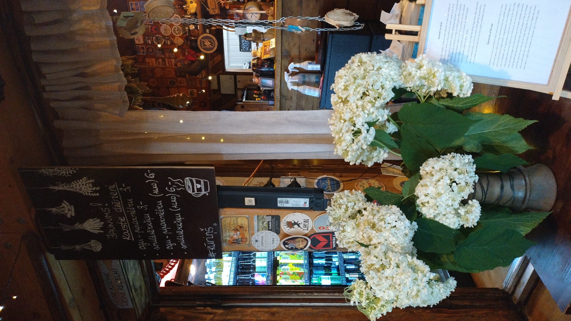 The image depicts the interior of a rustic establishment, possibly a cafe or bar, in Jūrmala, Latvia. In the foreground, a vase filled with large white hydrangeas sits on a wooden surface. To the left, a glass-fronted refrigerator is stocked with bottled beverages. A black chalkboard sign hangs prominently, displaying handwritten text and drawings in chalk. The sign lists "JAUNUMS!" (New!) followed by "Karstie dzērieni" (Hot drinks), with a menu of items and prices. Below the menu, a hashtag "#KŪRĪŅŠ" is written. Scattered on the wall behind the chalkboard are various circular stickers and patches. In the background, a counter area is visible with shelves and items on display, and a blurred individual can be seen. A cream-colored curtain hangs to the right of the chalkboard. The overall ambiance is cozy and informal, with natural wood elements and decorative touches.