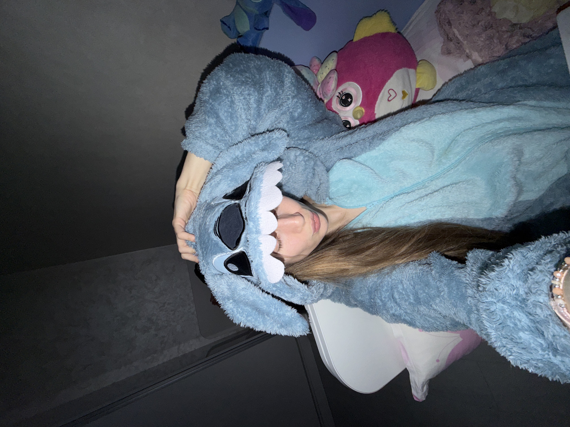 The image features a young woman lying down indoors, taking a selfie or being photographed from an overhead perspective. She is dressed in a soft, fuzzy blue kigurumi (onesie) designed to resemble the Disney character Stitch, with the character's face forming the hood around her head. Her long, light brown hair frames her face as she looks towards the camera with a neutral, relaxed expression, her lips slightly parted. She is using her left hand to hold the hood.
The setting appears to be a cozy indoor space, likely a bedroom, with several plush toys arranged around her. To her upper left, a smaller blue Stitch plush is visible. Directly above her head, a vibrant pink and yellow fish-like plush toy with large eyes and a white heart symbol on its cheek is prominently featured. Another fuzzy, lighter-colored plush toy with purple-brown hues is partially visible behind the pink plush. A white surface, possibly a pillow or a small table, is situated near her head. The background is predominantly dark grey or black, suggesting dim lighting, indicative of evening or nighttime. The overall atmosphere is one of comfort and leisure. The location context is Plovdiv, Bulgaria.