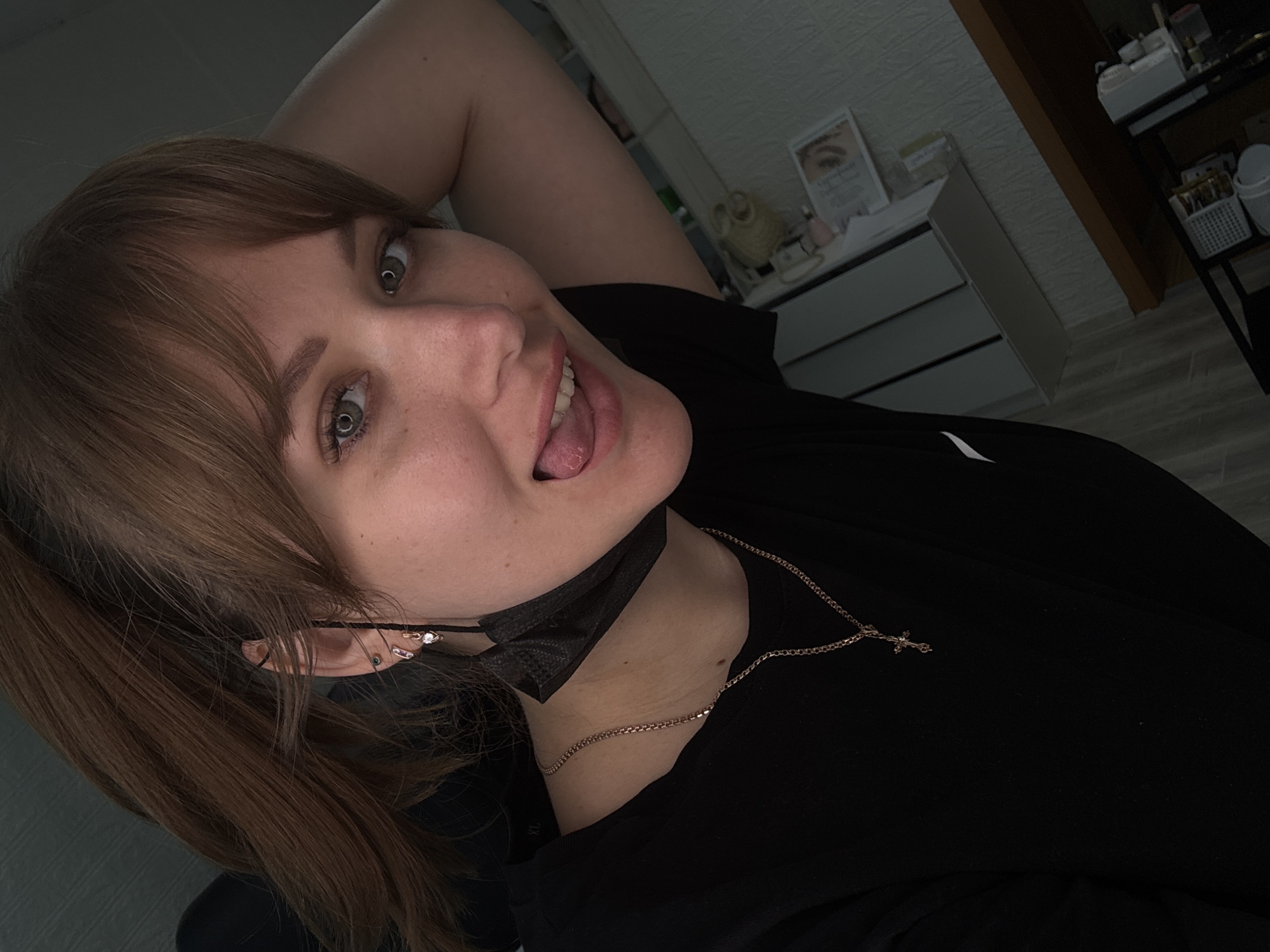 A young adult female is depicted in a close-up, chest-up indoor portrait, taken in Chaikovsky, Russia. Her reddish-brown hair, styled with bangs, falls over her forehead and around her face. Her green eyes are open and directed at the camera. Her complexion is fair, with visible freckles around her nose and cheeks. Her mouth is open, with her tongue extended past her lower lip in a playful expression. A black disposable face mask is pulled down below her chin, with one ear loop visible around her left ear.
She wears a black short-sleeved top. Around her neck are two gold-colored chains; a shorter, thinner chain and a longer, thicker chain from which a small cross pendant hangs on her chest. Her left ear (viewer's left) has multiple piercings: a small gold-colored hoop earring with a dangling heart-shaped charm (possibly with a small pink or clear stone) in the lobe, and a small stud earring in the upper cartilage. Her left arm is raised, with the elbow visible in the upper right quadrant of the image, suggesting her hand is behind her head or neck.
The background is an indoor setting. To the upper right, a white shelf unit or cabinet is visible against a light-colored, textured wall (possibly wallpaper). Below the shelf, a framed picture depicting a close-up of an eye with styled eyelashes is displayed. Further to the right and lower, a wooden surface and various white objects or containers are partially visible. The lighting is artificial and somewhat dim.