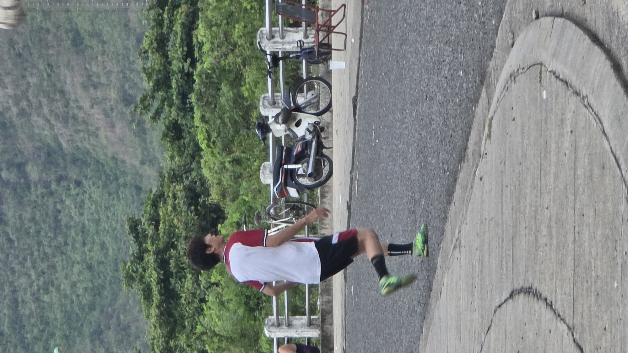 An outdoor, eye-level shot captures a young man in athletic attire engaged in physical activity on a paved path. The man, with dark, curly hair, wears a white t-shirt with red shoulders, black shorts, black athletic socks, and bright green athletic shoes. He is captured mid-movement, with his left leg extended forward and his body slightly tilted, suggesting he might be kicking or balancing.
To his left, a grey concrete barrier separates the path from a steep, densely vegetated green hillside or mountain range, stretching into the background. Behind this barrier, several vehicles are parked: two motorbikes (one white, one dark) and at least two bicycles. One of the bicycles is equipped with a red frame, possibly a child's seat or carrier.
The path he stands on is paved, possibly asphalt or concrete, and extends to the right foreground, where a distinct curved concrete edge borders it. The lighting appears soft and even, indicating it might be an overcast day or during the earlier/later hours of the day. There is no visible text in the image. The setting appears to be a scenic area, possibly a road or pathway overlooking a natural landscape in Bac Nha Trang Ward, Vietnam.