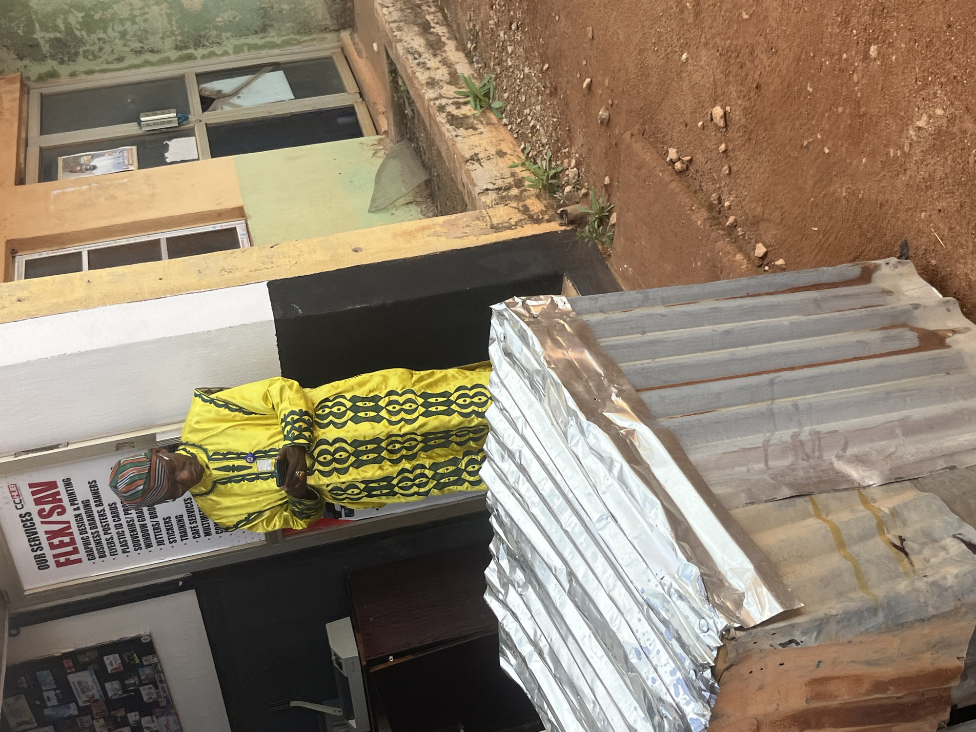In Gura Top, Nigeria, a man in a vibrant yellow garment with intricate green patterns stands near the entrance of a building. He wears a colorful striped cap and is looking down at his mobile phone, holding it with both hands. Behind him, a sign lists "OUR SERVICES FLEX/SAV" with bullet points detailing graphic design, printing, business branding, flyers, posters, banners, plastic ID cards, souvenirs, jotters, stickers, training, café services, and multimedia. To the left of the sign, a dark shelf unit is visible. The building's exterior features a mix of colors, including yellow, green, and pale orange plaster, with a glass door and a window. In the foreground, a structure covered in corrugated metal sheeting stands partially obscuring the view of the ground, which is composed of reddish-brown soil with scattered pebbles.