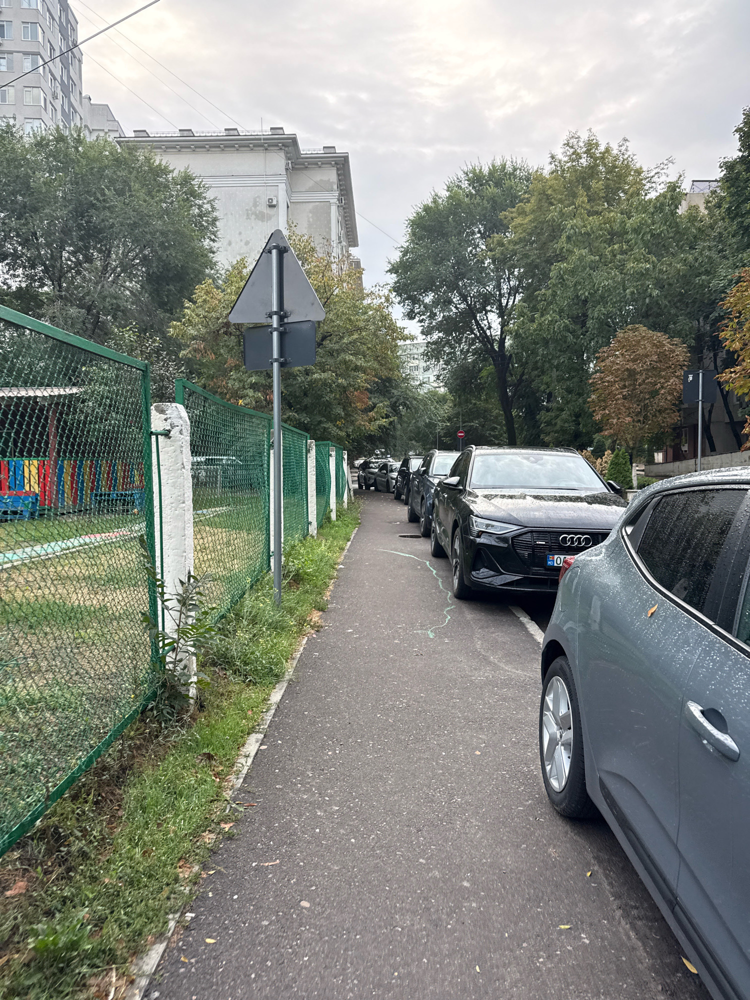 The image depicts a street scene in Chișinău, Moldova, on a cloudy day. A narrow asphalt sidewalk runs alongside a green chain-link fence. Beyond the fence, a playground area with colorful structures is visible. On the other side of the sidewalk, a row of parked cars lines the street. The cars are predominantly dark-colored SUVs. The foreground features the side of a gray car, its wheel and door handle visible. Further down the street, a black Audi e-tron is prominently parked. Trees with green and some autumnal foliage are present, casting shadows. A traffic sign, triangular and dark gray, is mounted on a pole next to the fence. Buildings and overhead power lines are also visible in the background. The asphalt appears damp, suggesting recent rain.
