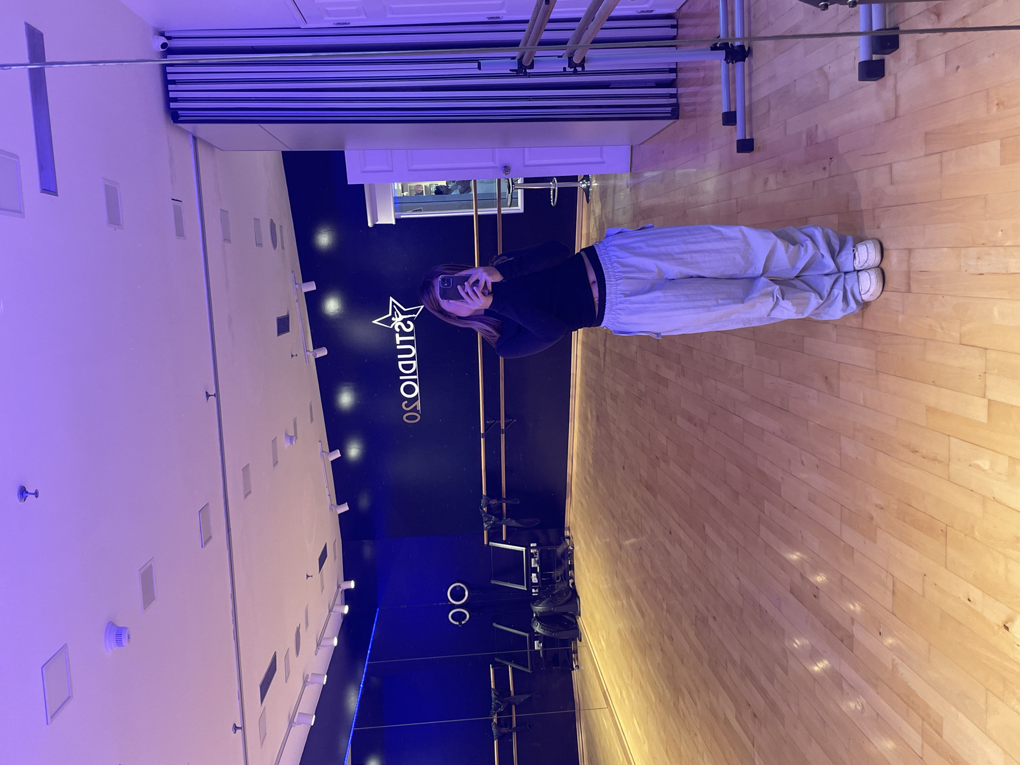 A person is standing in a dance studio, taking a selfie with their phone. They are wearing a black long-sleeved shirt and light-colored pants. The studio has a dark blue wall with a star logo and studio name on it. There are ballet barres along the wall. The floor is made of wood. Mirrors line the wall. The location in Porlamar, Venezuela is not discernible from this image.
