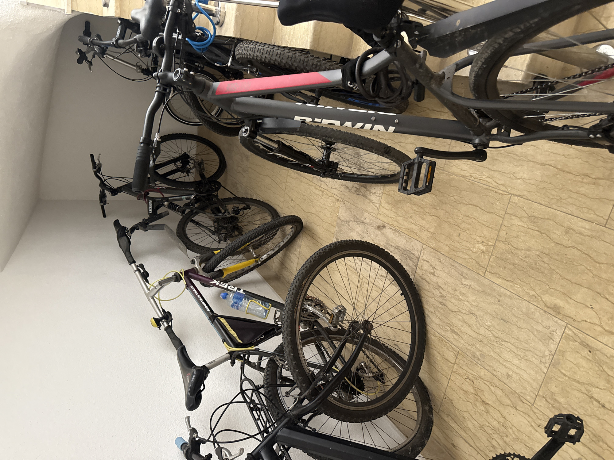 In Boreti, Montenegro, a collection of bicycles is stored in an indoor, sloped passageway. The passageway features off-white textured walls and a tiled floor with a light brown, marble-like pattern. The bicycles, varying in color from black to purple and yellow, are lined up against the wall, some propped against each other. Several are mountain bikes with knobby tires, suspension forks, and multiple gears. One bicycle in the foreground has a dark grey frame with pink and grey accents, and the brand name "BTWIN" is visible on its frame. Another bicycle, with a purple and yellow frame, has a bottle cage with a clear water bottle attached and the brand name "TREK" on its frame. The overall scene depicts a secure storage area for bicycles.