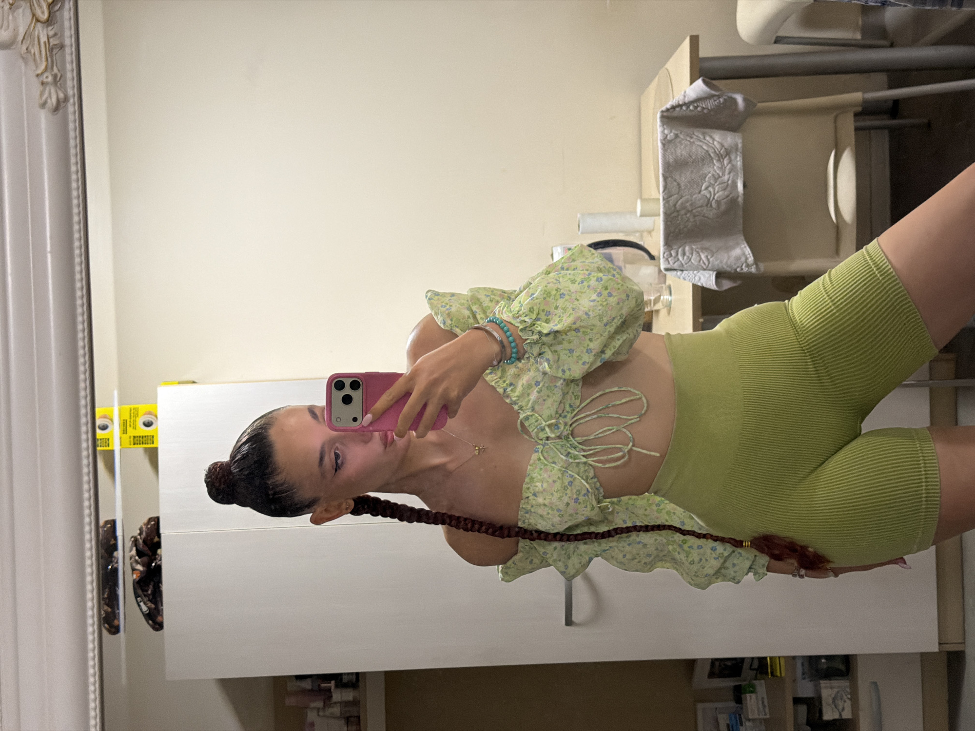 A young woman with dark hair styled in a top bun and a long braid takes a mirror selfie in what appears to be a residential interior, possibly a bedroom or bathroom, in Sofia, Bulgaria. She is wearing a light green floral off-the-shoulder crop top with puff sleeves and lime green ribbed high-waisted bike shorts. She is holding a pink smartphone in her right hand, angled to capture her reflection. A silver necklace with a small pendant is visible around her neck, and she has a bracelet on her left wrist. The background shows a white wardrobe to her left, and a table with a chair and a towel draped over it to her right. The overall lighting suggests an indoor setting, possibly illuminated by artificial light.