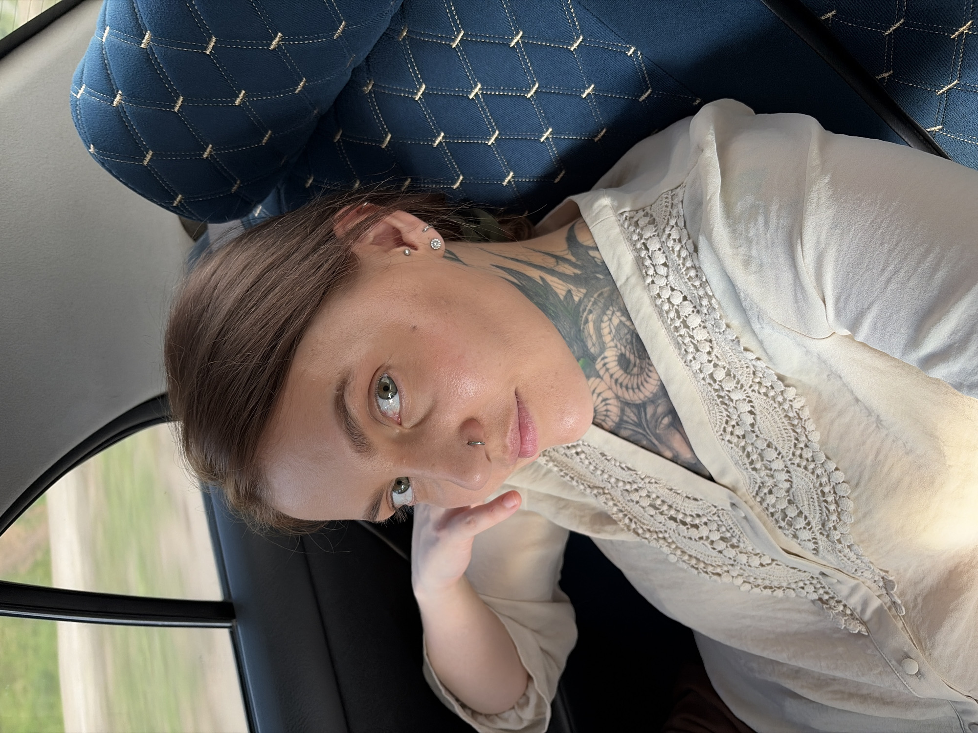 A young woman with light brown hair and striking green eyes is captured in a close-up, reclined shot, likely inside a vehicle, given the view of blurred greenery and a road through the window. She is wearing a light-colored, button-down shirt with intricate lace detailing along the collar and placket. Her visible skin shows a large, intricate tattoo on her neck and upper chest. She has piercings in her ear and nose, and her expression is relaxed and somewhat contemplative. The background reveals the quilted upholstery of a vehicle's seat, in a deep blue hue. The lighting suggests it is daytime, possibly late afternoon given the warm tones. The image is slightly tilted, adding a dynamic perspective to the portrait.