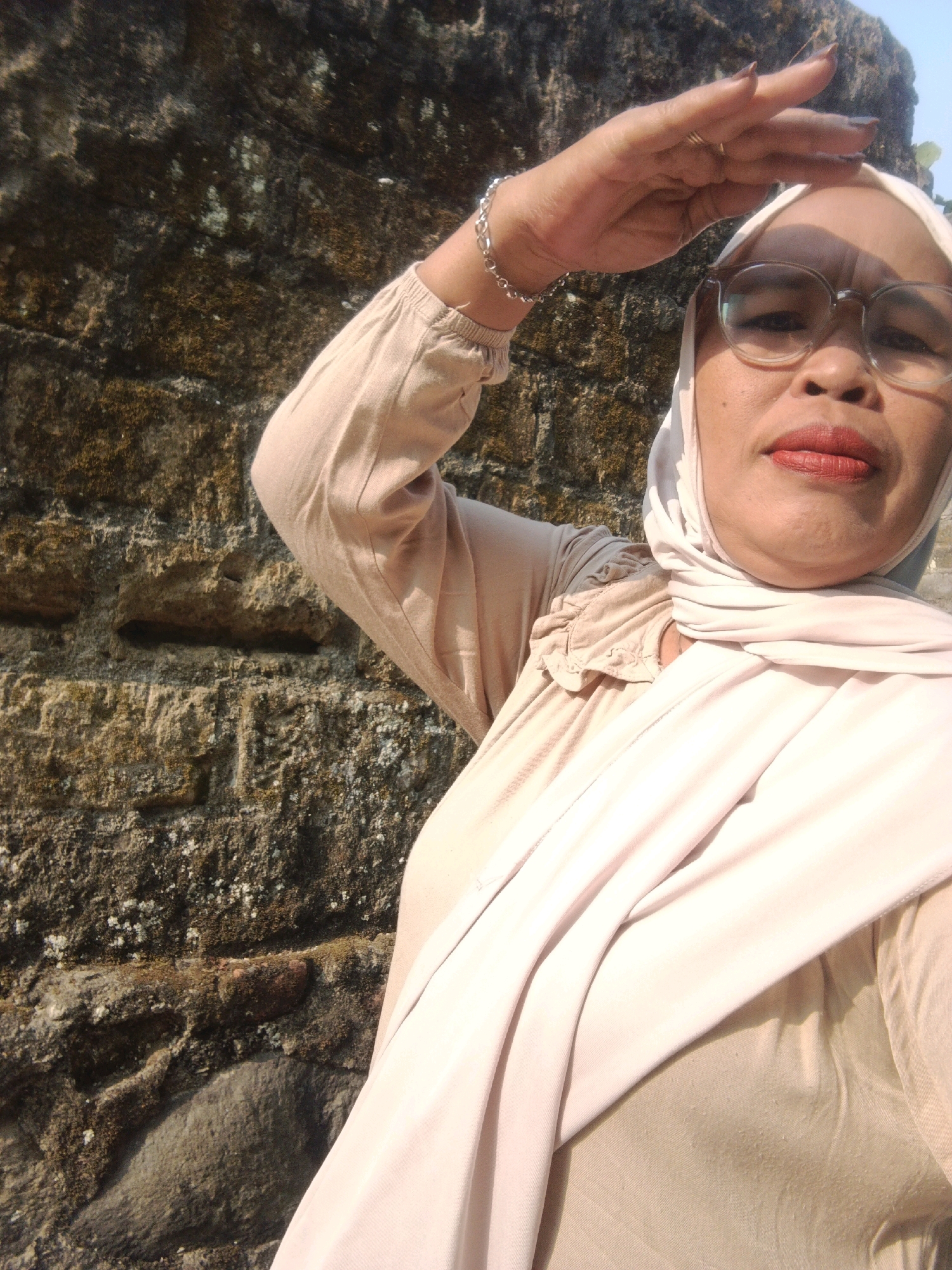 A woman wearing a hijab and glasses is taking a selfie outdoors, with a stone wall in the background. The woman is in the foreground, obscuring most of the wall. She is holding her right hand up to her forehead, shielding her eyes from the sun. She is wearing a light-colored hijab and a matching long-sleeved shirt. The stone wall appears to be made of rough-hewn blocks, some of which are covered in moss. The lighting suggests it is daytime. The location is identified as Warungkiara, Indonesia.