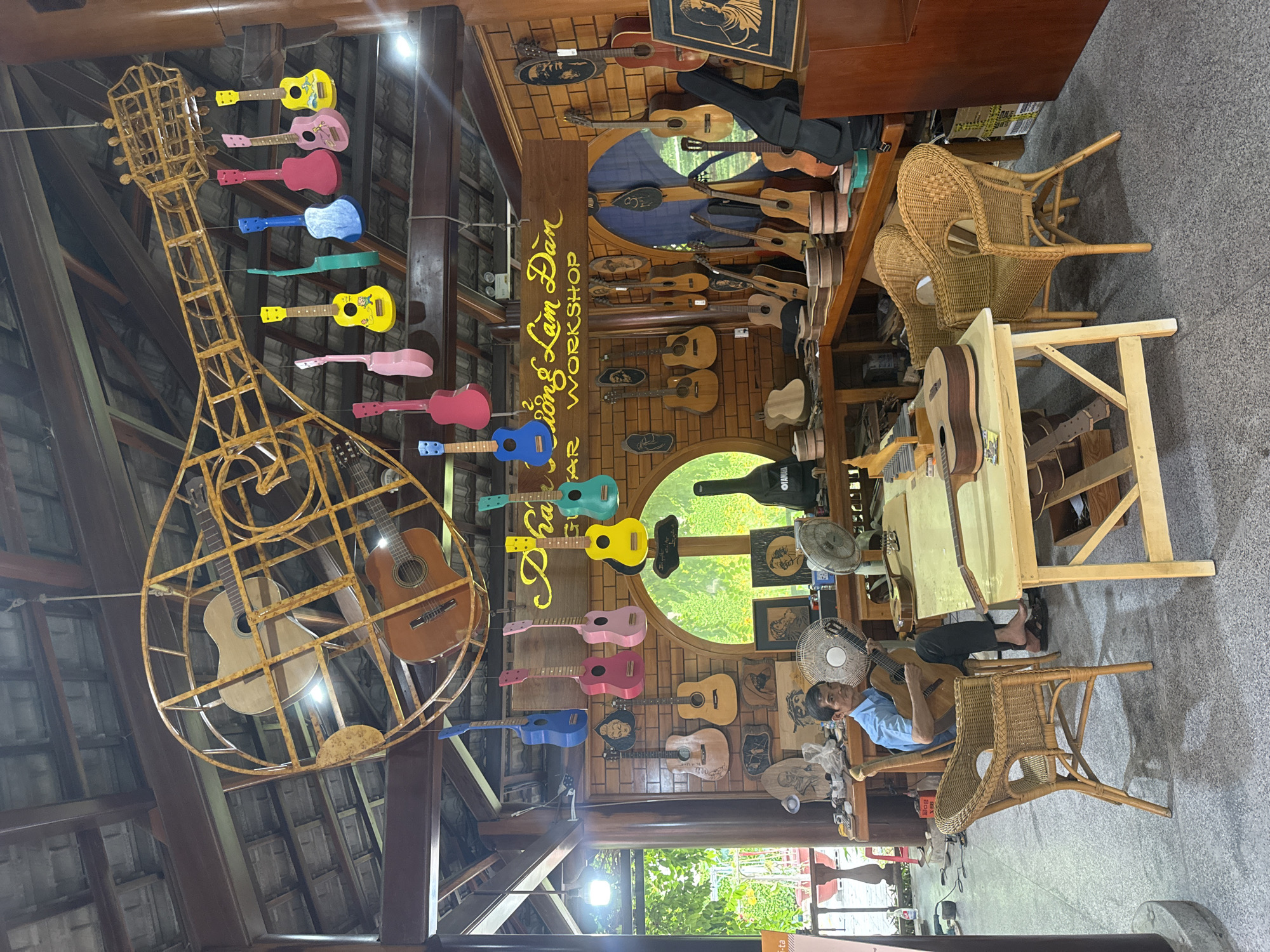 The image depicts the interior of a guitar workshop in Nha Trang, Vietnam, specifically the "Phuong Lam Dan Guitar Workshop". The workshop is filled with guitars of various sizes and colors, some hanging from the ceiling and others displayed on walls and shelves. In the foreground, a man is seated on a wicker chair, holding and playing a guitar. He is wearing a blue shirt and dark pants. Two wooden tables are present, one with a guitar lying on it, and the other with a guitar being played by the man. Wicker chairs are placed around the tables. The walls are constructed of brick, and a wooden beam ceiling with exposed rafters is visible. Decorative elements include a large metal sculpture of a guitar suspended from the ceiling, numerous smaller guitars hanging at different heights, and framed artwork on the walls. A fan is visible near the seated man, and other fans and musical equipment are scattered throughout the workshop. The overall ambiance suggests a working environment dedicated to the craft of guitar making and playing.