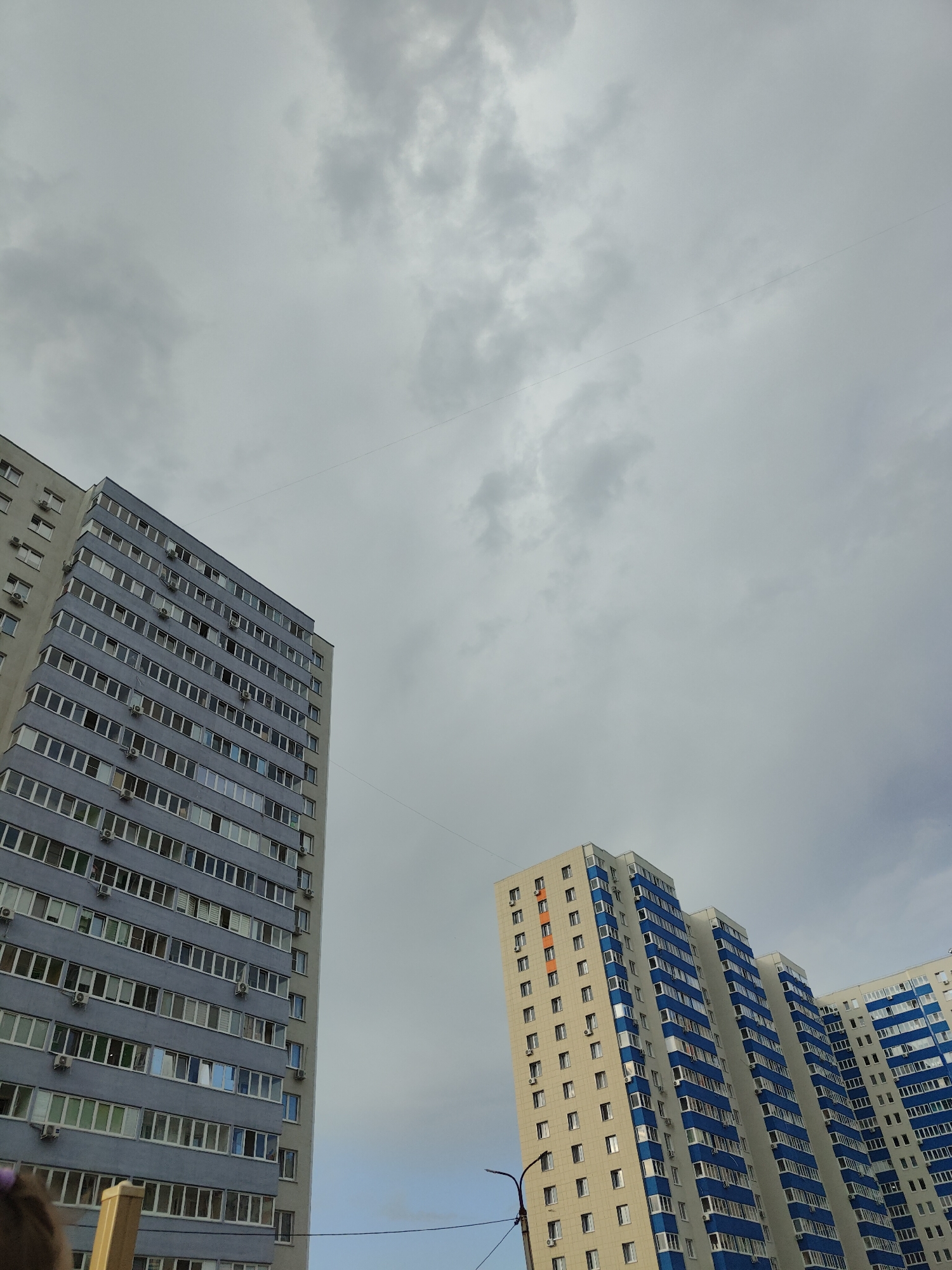 The image captures a view of several high-rise residential buildings against a cloudy sky. Two prominent buildings are in focus, viewed from a low angle looking upwards. The building on the left is tall and appears to be light gray with numerous windows, some of which are tinted blue. Air conditioning units are visible mounted outside many of the windows. The building on the right is a cluster of connected towers, primarily cream-colored with striking blue accents around the balconies and window frames. It also features many windows and air conditioning units. A thin, dark line, likely a wire, stretches diagonally across the sky. Another building, mostly obscured by the foreground structure on the right, is also visible. A street lamp is partially visible in the lower right portion of the image. The sky is overcast with varying shades of gray clouds, suggesting a cloudy or possibly rainy day. No people are clearly discernible, though a small, dark object, possibly part of a person's head or clothing, is visible in the bottom left corner. The overall scene suggests a residential urban environment in Ufa, Russia, likely during the daytime. There is no visible text in the image.