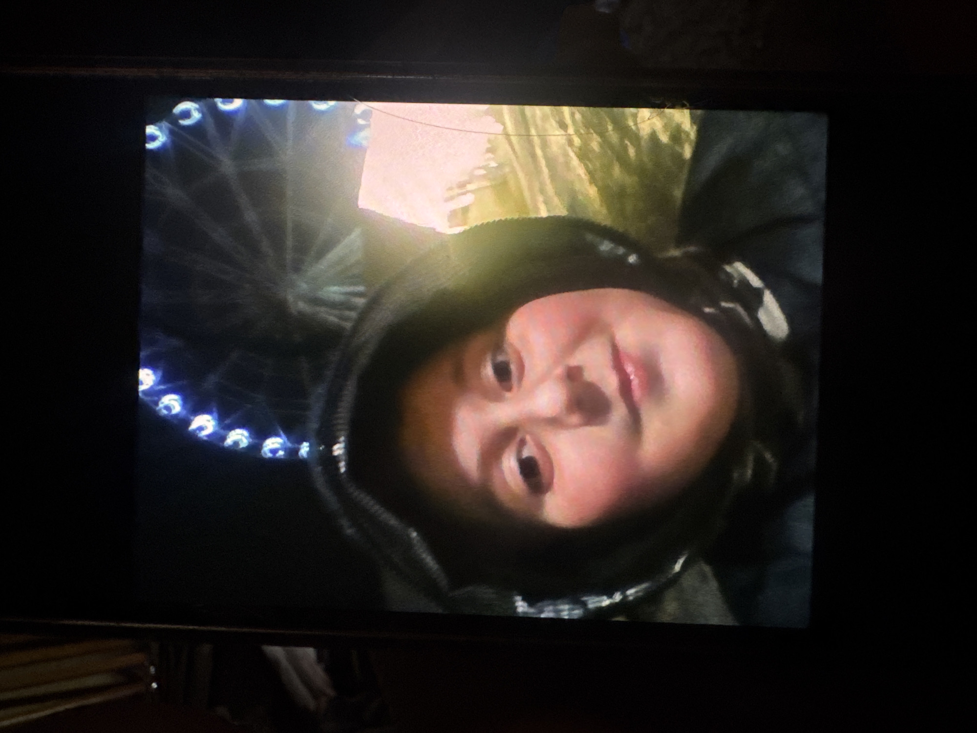 The media file captures a close-up, slightly tilted shot of a young child wearing a dark hooded jacket, looking directly at the camera. The child has rosy cheeks and a broad smile. In the background, a Ferris wheel with illuminated blue lights is visible, suggesting the image was taken at a nighttime event, likely an amusement park or fair. The overall lighting is dim, with a glow emanating from the Ferris wheel and possibly a light source illuminating the child's face. The scene appears to be outdoors, given the presence of the Ferris wheel and the dark, possibly natural, background elements. The child's expression conveys happiness and a playful mood. There is no visible text in the image. The location context provided is Rostov-on-Don, Russia.