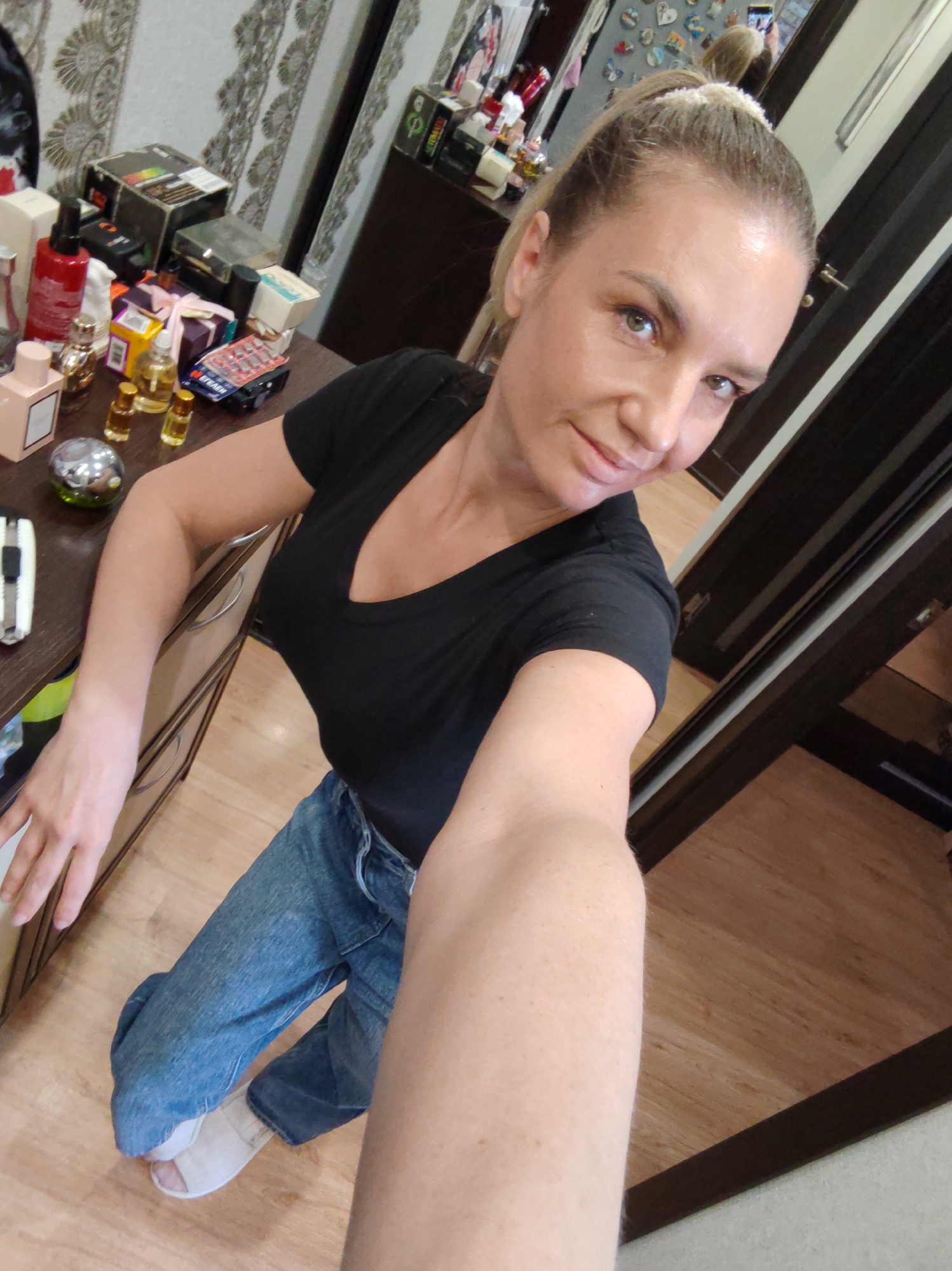 A female individual is depicted in a residential interior in Petropavlovsk-Kamchatsky, Russia, taking a selfie. Her blonde hair is styled in a high ponytail secured with a light-colored scrunchie. She has light skin and displays visible skin texture. She wears a black V-neck t-shirt, blue denim jeans, and light-colored open-toed slip-on shoes. Her greenish-brown eyes are directed at the camera, and a subtle smile is present. Her right arm is extended, holding the camera for the photograph, while her left hand rests on the surface of a dark brown wooden dresser.
The dresser's surface is extensively covered with various items, including multiple fragrance bottles such as a pink rectangular bottle and several small amber-colored vials, a red pump bottle, and a spherical silver object with green visible beneath. Other objects include a white-handled utility knife, a black package labeled "STECH" containing blister packs, a white rectangular box, and a purple-wrapped item secured with a pink ribbon.
The background features a wall adorned with light-colored, ornate, floral-patterned wallpaper. Behind the subject, a large mirror reflects parts of the room, including a surface decorated with numerous small, multi-shaped magnets, as well as the top of the subject's head and the camera device. To the right, dark brown wooden door frames or closet elements with a silver handle are visible. The flooring throughout the visible area is light brown wood laminate or parquet. The individual is positioned in a crouched or kneeling posture.
