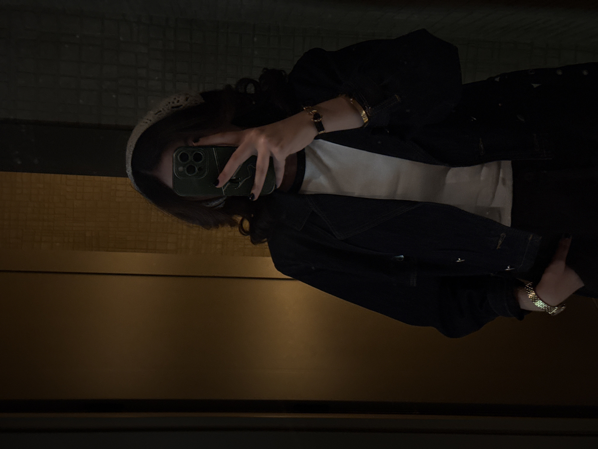 A mirror selfie captures a person from the chest up, holding a dark green smartphone that obscures their face. The person is wearing a dark denim jacket over a white top, with dark hair and a light-colored, possibly crocheted, headband. Their hands and wrists are visible; one wrist is adorned with a gold-toned bracelet and a wristwatch featuring a black band, while the other displays a metal-link wristwatch. Dark nail polish is visible on the fingers holding the phone. The reflected background, indicating an indoor setting in Yerevan, Armenia, shows a dark, possibly tiled wall in the upper portion and a warm, golden-brown horizontal panel below. The dim lighting suggests an evening or a low-lit interior. No text is visible in the image.