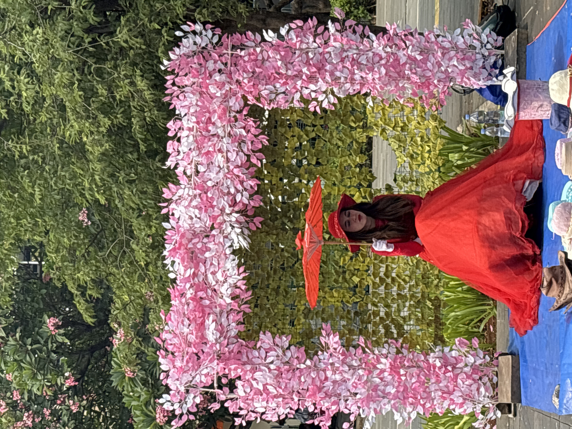 A woman in a red dress and matching hat, holding a red umbrella, sits on a blue tarp in front of a green hedge wall and a floral archway made of pink and white artificial leaves. The setting appears to be an outdoor public space, possibly a park or a street market, in West Jakarta, Indonesia, as suggested by the greenery and the overall atmosphere. Several hats are arranged on the blue tarp in front of the woman, some of which appear to be for sale or display. Two bottles of water are placed behind her on the ground. The woman's face is partially obscured by her hat and umbrella, but she appears to be looking down. The scene is illuminated by natural daylight, casting soft shadows.
