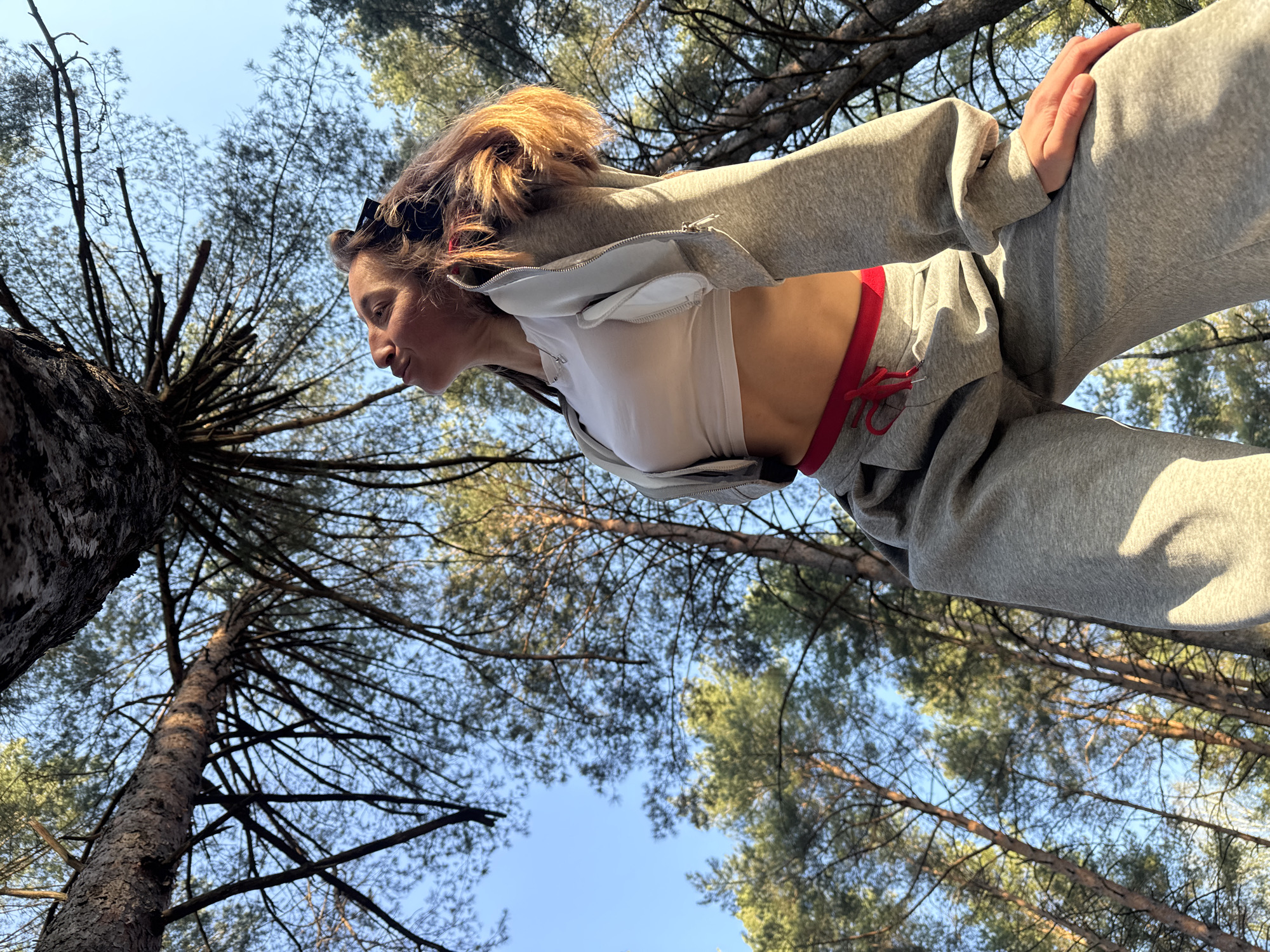 A low-angle shot depicts a woman against a backdrop of trees and a clear blue sky. The woman is wearing a white top and a grey tracksuit with red accents. She appears to be standing, with her hand resting on her thigh. The trees, presumably in a forest or wooded area, stretch upwards towards the sky. The location is in Прикамский, Russia.
