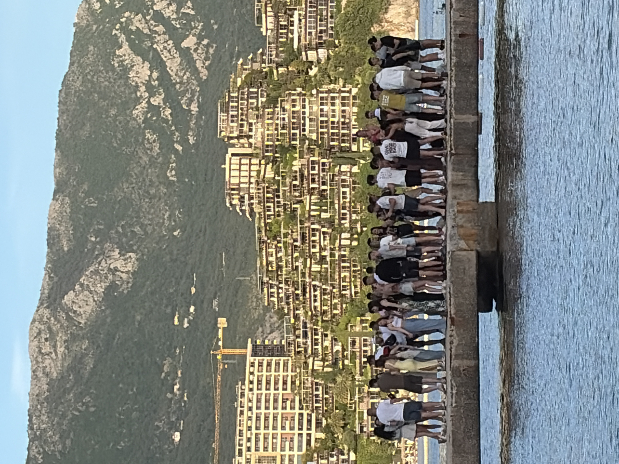 A scenic view shows a group of people standing on a concrete pier, likely tourists, facing the coastal city of Budva, Montenegro. In the background, the landscape features mountains, and buildings, giving the impression of a densely built environment. A construction crane is also visible. The pier extends into the water to the right. The scene is observed in daylight with natural lighting illuminating the architecture, the group of people, and the surrounding area.
