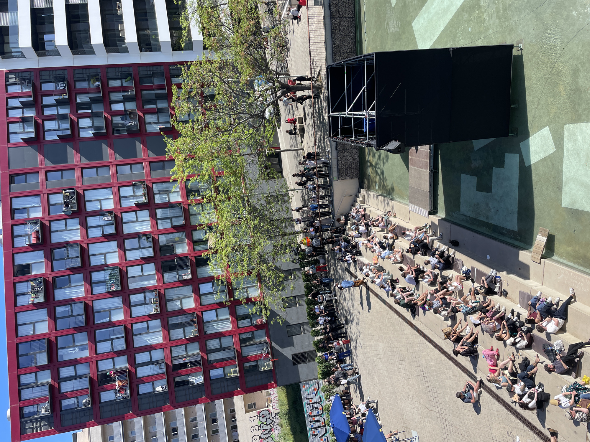 The image depicts an outdoor gathering of people in a city square. A modern red building with many windows stands on the left, contrasted by a taller white building in the background. In the center, a tree with green leaves provides partial shade. A black structure, potentially a stage or screen, is positioned in front of a water feature. The audience is seated on stone tiers, watching a performance, relaxing, or sunbathing. Several people are standing near the stage or on a walkway behind the seated audience. The area appears to be well-maintained, with clean pavements and minimal clutter. The image likely captures an event or performance taking place in a public urban space in Barcelona, Spain.