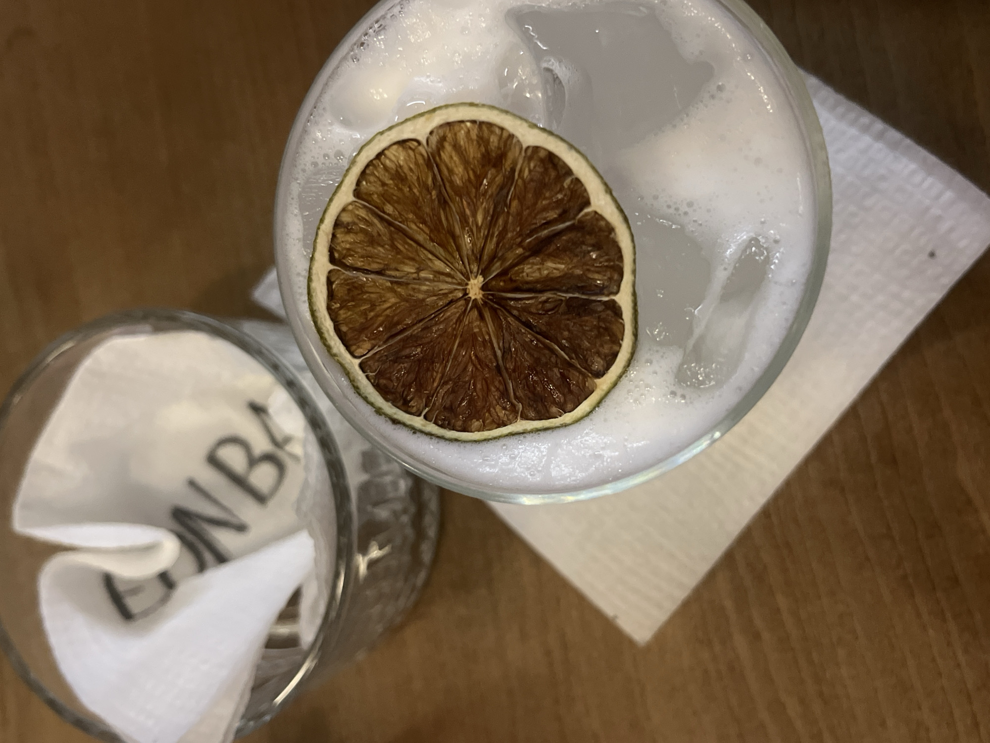 The image is a high-angle, still-life shot of two drinks on a wooden surface, likely a table. The drink on the right is in a clear glass, appears to be a cocktail with a frothy top, and is garnished with a dehydrated lime slice. The drink sits atop a white napkin. To the left is another clear glass containing a white napkin, possibly crumpled or used. The background consists of a wooden surface with a distinct grain pattern. The setting appears to be a bar or restaurant in Sofia, Bulgaria, based on the "Bon Bar" inscription visible on the napkin in the second glass. The lighting is somewhat dim, suggesting an indoor environment.