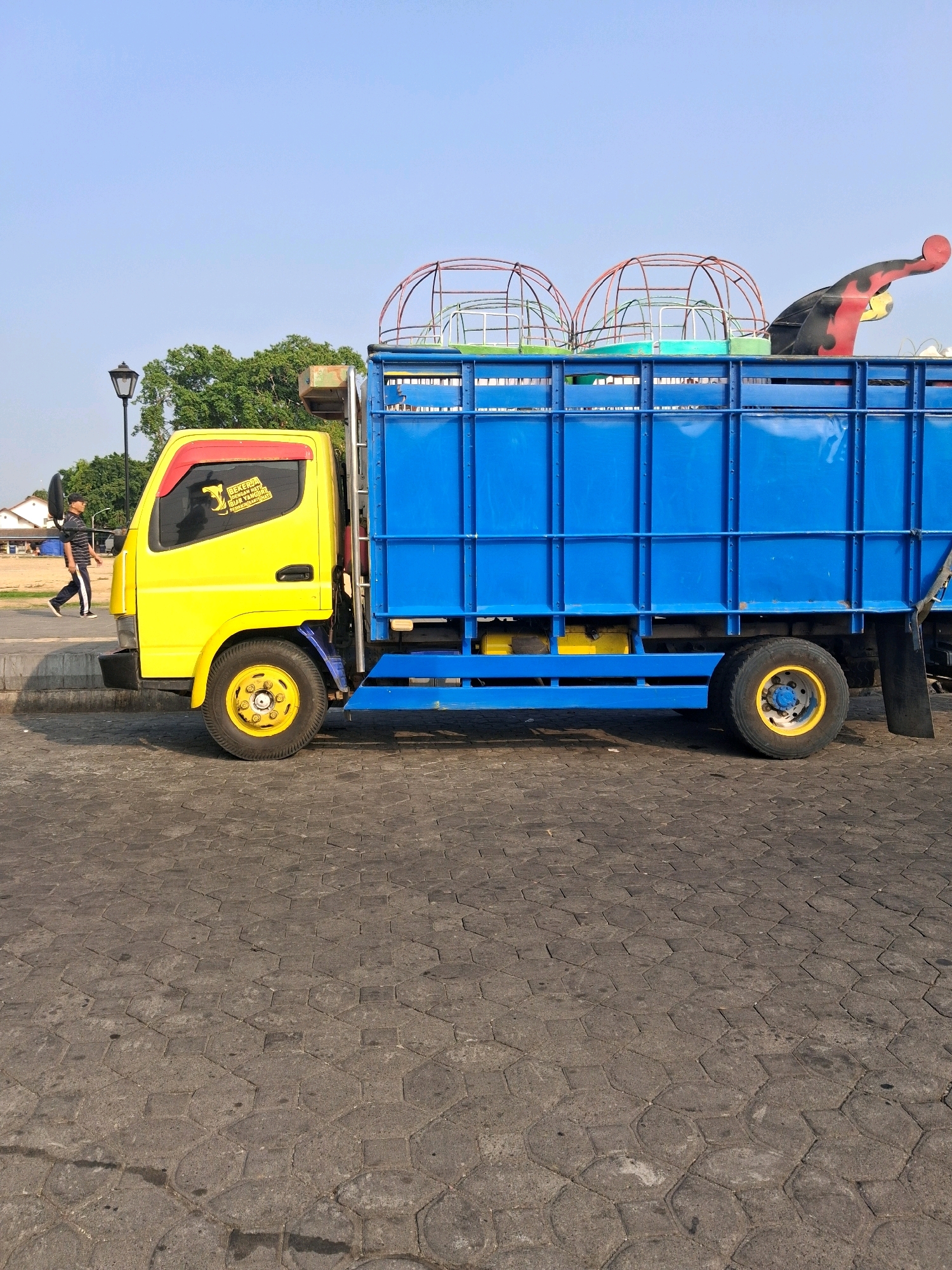 In Surakarta, Indonesia, a yellow and blue truck is parked on a cobblestone street. The truck's cab is yellow with red trim around the windows, and the cargo bed is bright blue. On the side of the cab, a sticker with text and a snake graphic is visible. The truck is loaded with what appear to be disassembled parts of a carnival ride, including a red and brown metal structure resembling a dome and a colorful, stylized dragon or sea creature statue. The street is paved with hexagonal grey cobblestones. In the background, a person wearing a black and white striped shirt and dark pants walks away from the truck, and a lamppost and green trees are visible. The sky is clear and blue.