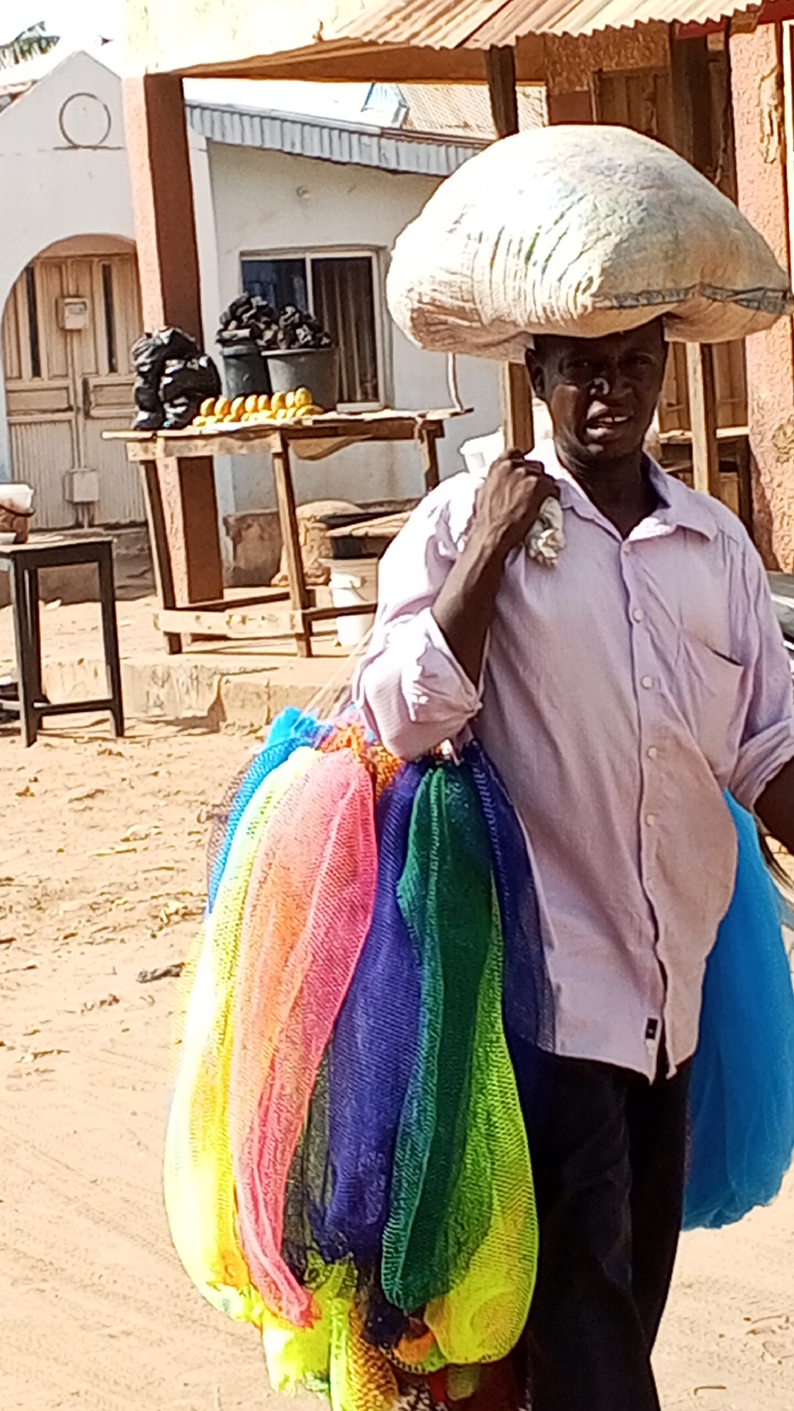 A man is carrying a large bag on his head and a bunch of colorful mesh bags on his shoulder in Sabon Tasha, Nigeria. The man is wearing a light pink, long-sleeved shirt and dark pants. The bags on his shoulder are a rainbow of colors, including yellow, pink, blue, green, and orange. Behind him, there is a table with items that appear to be charcoal, bananas, and other objects for sale. There is a building in the background. The ground is dirt.