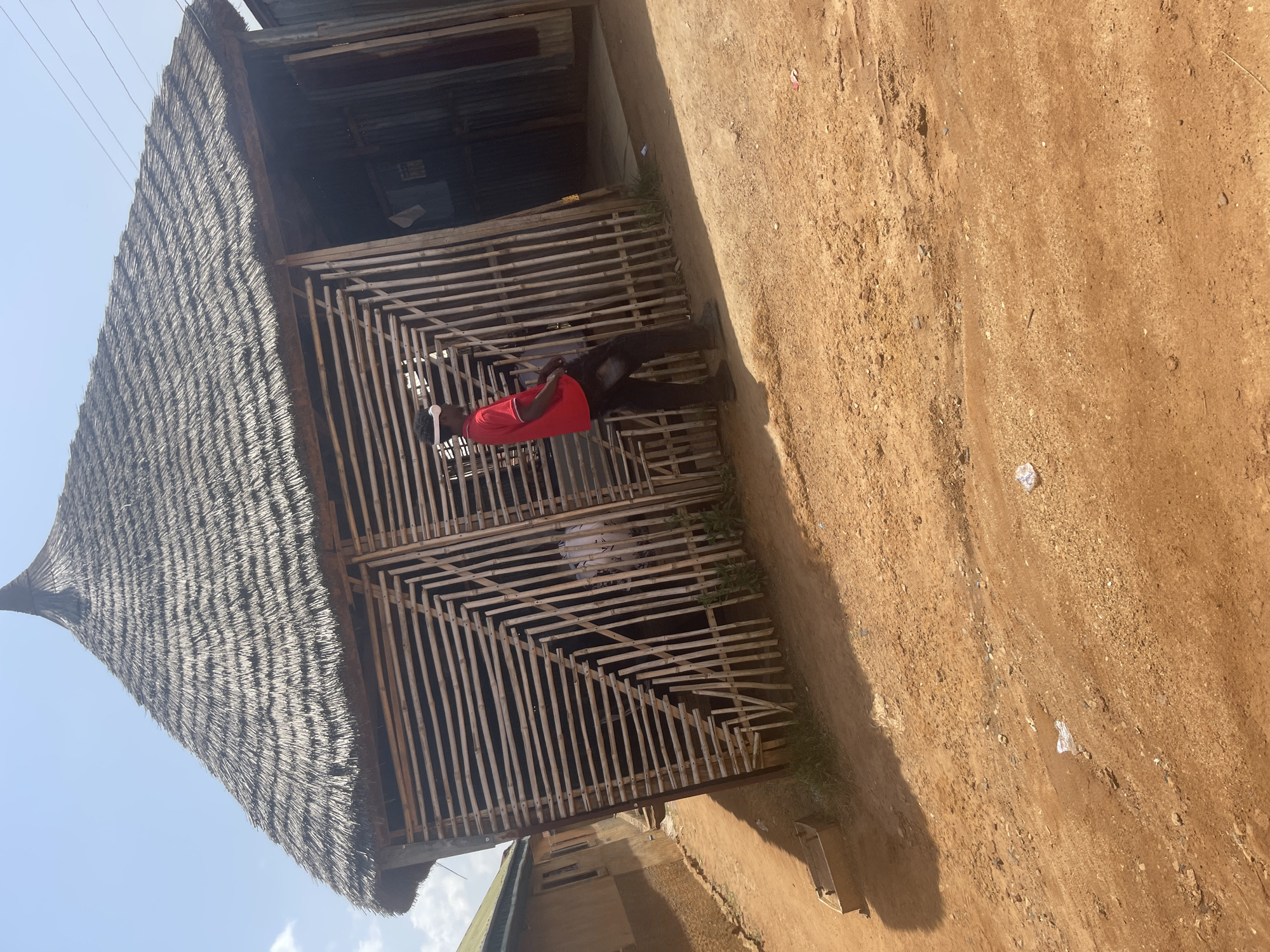 A person in a red shirt and black pants stands in front of a thatched-roof structure with bamboo walls. The structure appears to be a shop or open-air stall. The ground is made of dry, reddish-brown dirt with scattered small rocks and some plastic litter. The sky is visible in the upper left corner, with some clouds and power lines. The scene suggests a rural or semi-rural location in Nigeria, possibly during daylight hours. The person appears to be looking down or at something to their side. The overall impression is one of everyday life in a warm climate.