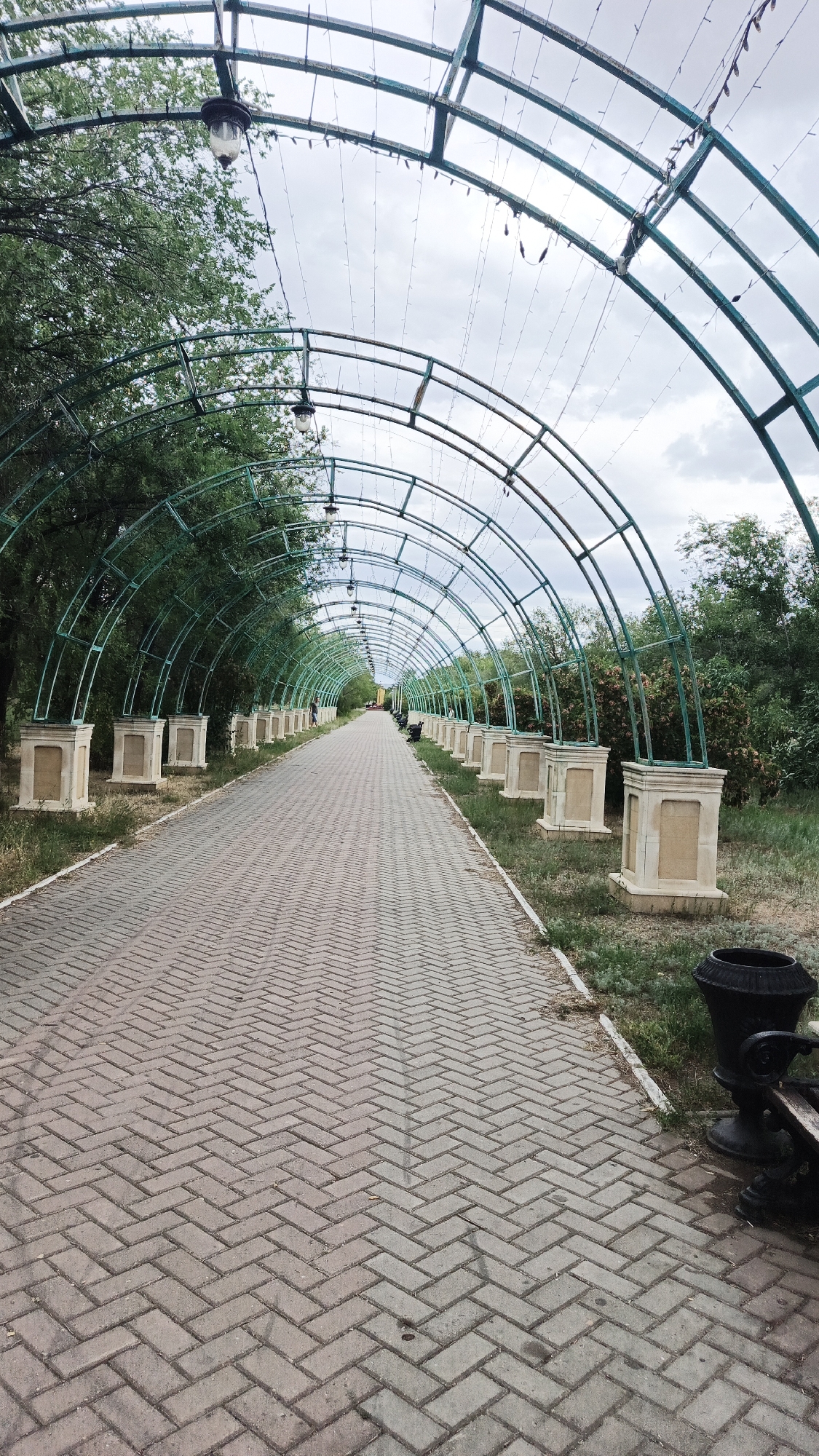 The image depicts a long, arched pathway in a park in Aksay, Kazakhstan. The pathway is paved with herringbone-patterned bricks and is lined on both sides by a series of arched, green metal structures that form a tunnel-like effect. These structures are supported by decorative, light-colored bases and appear to have string lights and hanging lamps attached. Trees and greenery flank the pathway, creating a natural setting. In the distance, a single person is visible walking along the path, and there are benches and a trash receptacle near the foreground on the right side. The sky is overcast.