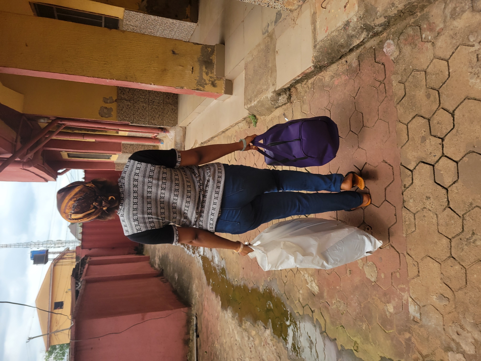 The image shows a person walking on a paved walkway with a purple duffel bag in their left hand and a white plastic bag in their right hand. The person is wearing a patterned long-sleeved shirt and blue jeans. Their back is to the camera, and they are wearing orange sandals. The walkway is made of hexagonal pavers, and there are some wet patches on the ground, suggesting recent rain or a spill.
In the background, there are buildings with yellow and red facades. The architecture appears to be residential or commercial, typical of a neighborhood in Nigeria. One building has a textured yellow wall with pink trim. Another section shows a red corrugated metal fence. Above the buildings, a cloudy sky is visible. The overall scene suggests an outdoor environment in a town or city.
The time of day appears to be daytime, possibly late morning or afternoon, given the lighting. There are no discernible emotions visible as the person's face is not shown. The activity is simple: walking with belongings, perhaps returning from or heading to a destination. The location context of Egbagu, Nigeria, aligns with the visual cues of the architecture and the paved outdoor setting.