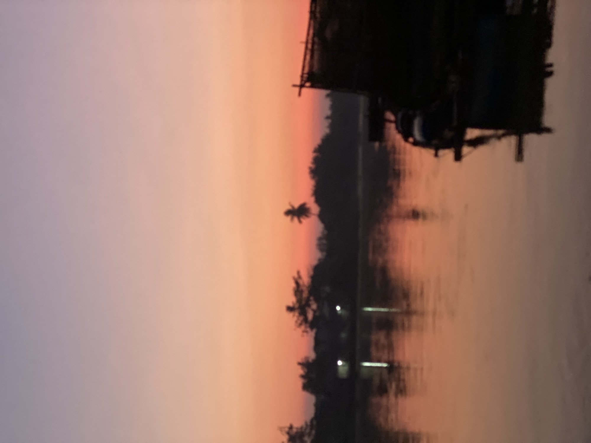 The image depicts a blurry, silhouetted view of a canal in Banjarmasin, Indonesia, during twilight. The sky displays a gradient of soft purples and oranges, transitioning from a pale lilac at the top to a warm peach and pink closer to the horizon.
The water surface of the canal reflects the sky's colors, creating a smooth, shimmering expanse. On the far bank, a dark, silhouetted line of trees and vegetation is visible, with a few faint lights indicating buildings or settlements. A distinct palm tree silhouette stands out against the horizon.
In the foreground and to the right of the frame, the dark silhouette of a boat or a floating structure is prominent. The structure appears to have a slatted or open-sided portion on top, possibly a shelter or a fishing net enclosure. The reflection of this structure is visible in the water below. The overall impression is one of tranquility and the muted beauty of a tropical evening.