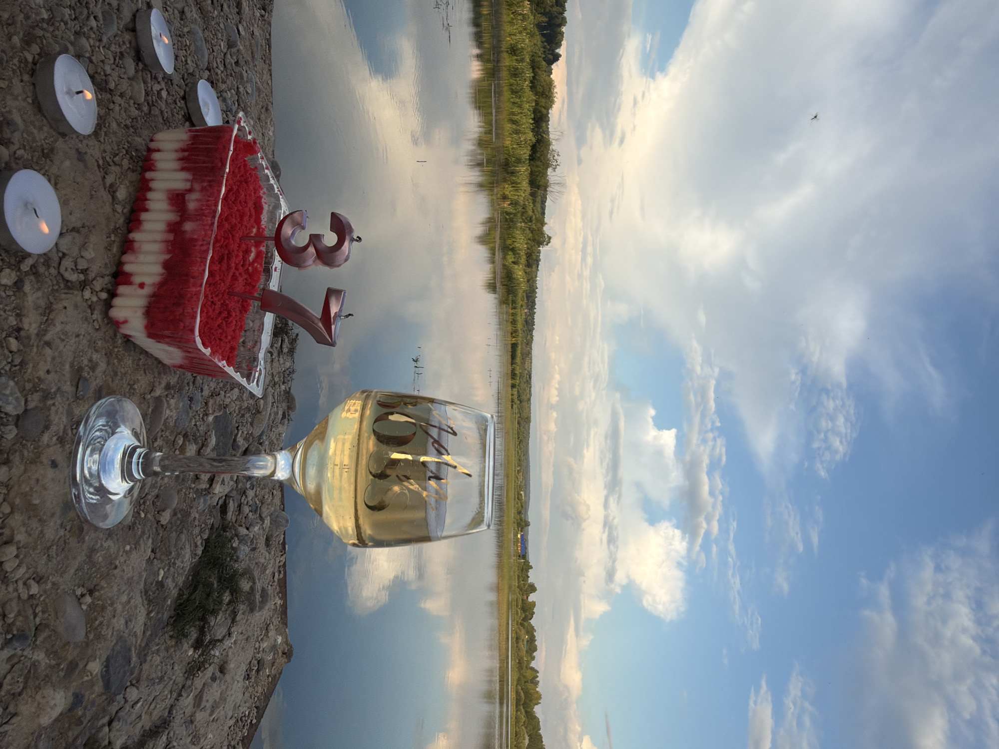 The image depicts a still life scene by a body of water, likely a lake or river, on a cloudy day. In the foreground, on a rocky surface, are a plastic container with a dessert topped with red sprinkles and white cream, two rose gold number candles forming "37", and four lit tea light candles. To the right of the dessert, a wine glass filled with a light-colored liquid, possibly white wine or champagne, is placed. The glass has gold lettering that appears to read "Jolly Boss". The background features a calm body of water reflecting the sky, with a line of trees and vegetation along the far bank. The sky is a mix of blue and white clouds. There is no indication of the city of Bolshaya Usa.