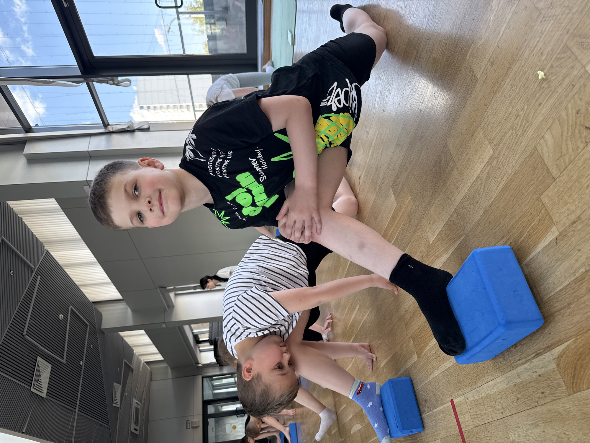 The image shows two young boys participating in a physical activity, likely a stretching or yoga class, held indoors in Khimki, Russia. The setting is a brightly lit room with a light-colored wooden floor and large windows revealing a clear blue sky with white clouds, indicating daytime.
In the foreground, a boy with short brown hair, wearing a black t-shirt and black shorts, looks directly at the camera with a subtle smile. His t-shirt is printed with green graphics and text, including "POSITIVEND / POSITIVE / POSITIVE LIE" and "Summer Holiday," along with the word "SUPER." He is in a lunge-like position, with his left foot elevated on a blue yoga block. Adjacent to him, another boy with short brown hair, wearing a black and white striped t-shirt and dark pants, performs a similar stretch. He is bent forward with his hands on the floor, and his right foot rests on another blue yoga block. His socks are blue with white stars and feature a small red bus or van design.
Several other individuals are visible in the background, suggesting a group class or training session. A small, light-yellow piece of popcorn or a similar item is noticeable on the wooden floor.