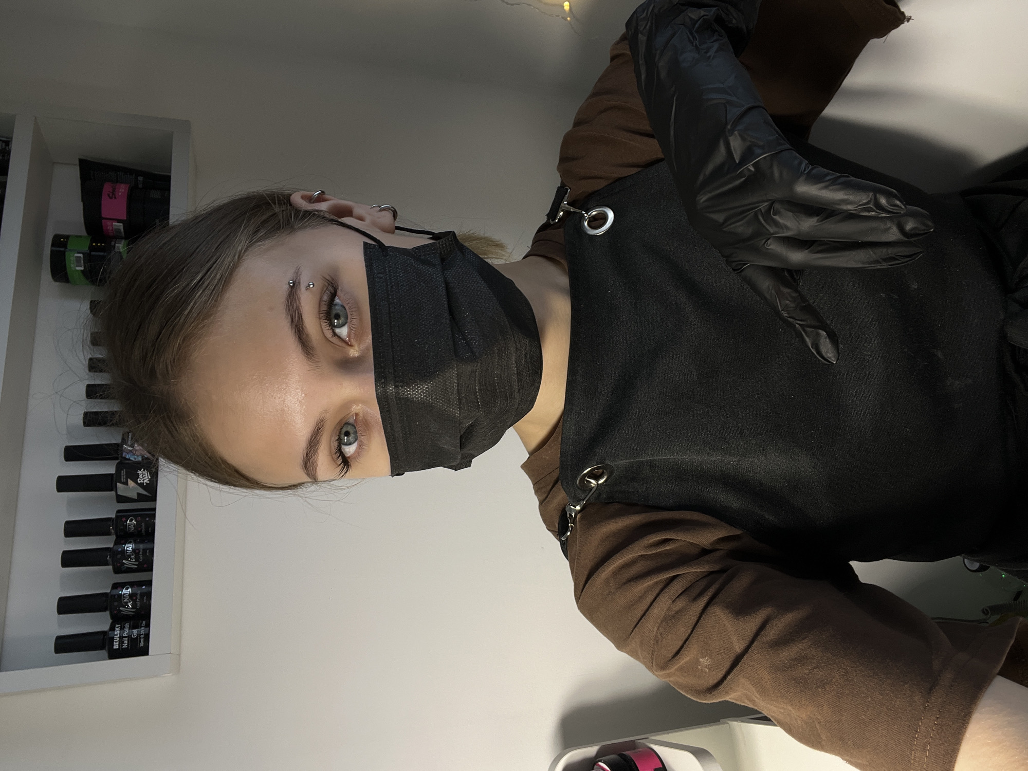A young person wearing a black face mask and black gloves, likely a manicurist or beautician, is positioned in front of a white wall. The person is wearing a brown long-sleeved shirt under a black apron. Their left eyebrow has a silver piercing. Behind them to the left, there's a shelf with bottles of nail polish. The setting appears to be an indoor salon or studio. There is no verifiable evidence to link this image to the city of Фоки, Russia.