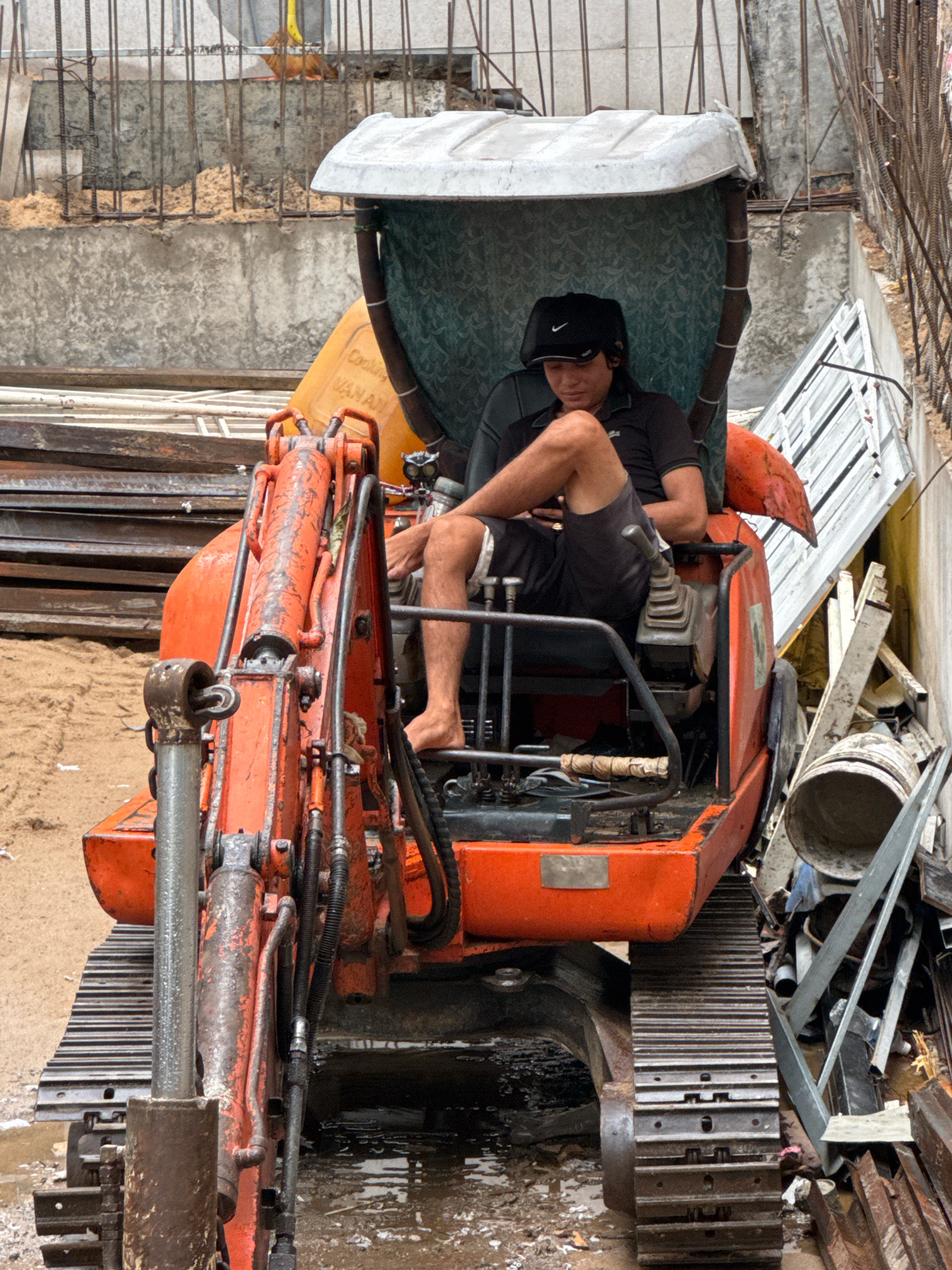 A young male construction worker wearing a black cap and a black polo shirt is seated in the driver's seat of an orange excavator on a construction site. He is barefoot and wearing black shorts. The excavator is orange and has a green fabric covering over the top. In the background, there are concrete walls, rebar, construction materials, and piles of dirt. The ground is muddy and wet. The scene appears to be outdoors in a developing area. The specific location is a construction site in the city of Nha Trang, Vietnam.