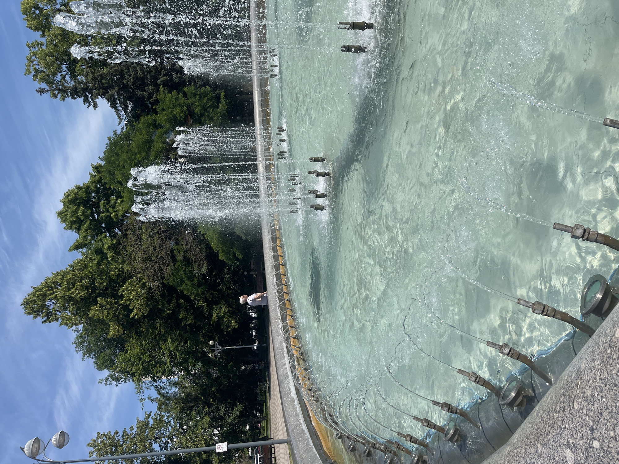 The image depicts a close-up view of a fountain in a park, likely located in Krasnodar, Russia. The fountain's water is a clear, light turquoise color, with numerous jets of water shooting upwards from several nozzles along the edge and from submerged spouts within the basin. In the background, a person wearing a white shirt and cap is seen walking along a curved pathway. The park features mature trees with dense green foliage and a bright blue sky with wispy white clouds overhead. A streetlamp with two globes is visible on the left side of the image, near a sign. The overall scene suggests a sunny day in a public park setting.