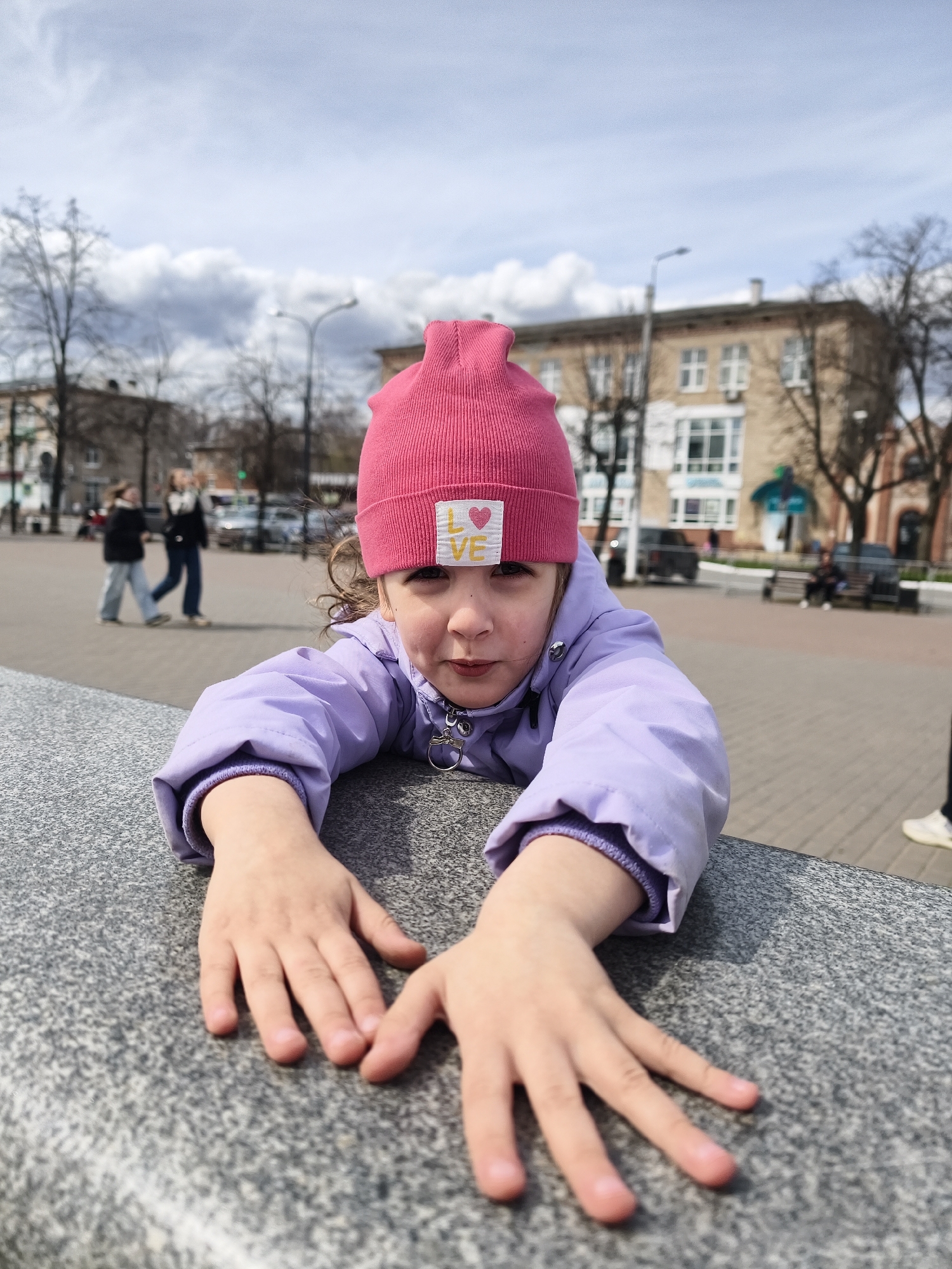 A young girl wearing a pink beanie with a "LOVE" patch and a purple jacket is leaning over a gray stone wall. She extends both hands toward the camera. In the background is a city square with sparse trees. A building is visible behind her, and people can be seen walking. This photo was taken in the city of Chaikovsky, Russia.