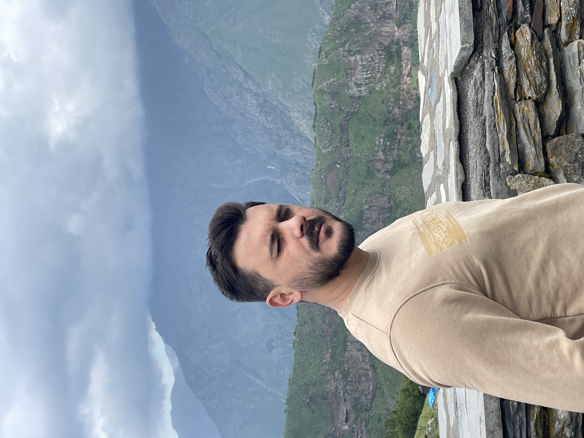 A man with a beard and dark hair stands in the foreground with his eyes closed, facing slightly to the right. He is wearing a light brown sweatshirt with a golden rectangular design on the left chest. Behind him, a stone wall or barrier is visible, constructed from irregular gray and brown stones. Beyond the wall, a lush green mountainside is illuminated by daylight, with a dramatic, cloudy sky overhead. The distant mountains are shrouded in a hazy, atmospheric perspective. The overall scene suggests an outdoor location, likely in a mountainous region, with hints of a remote or natural setting. The presence of a blue tarp and a fence on the left side of the frame suggest a possible man-made structure or a viewing area in the landscape.