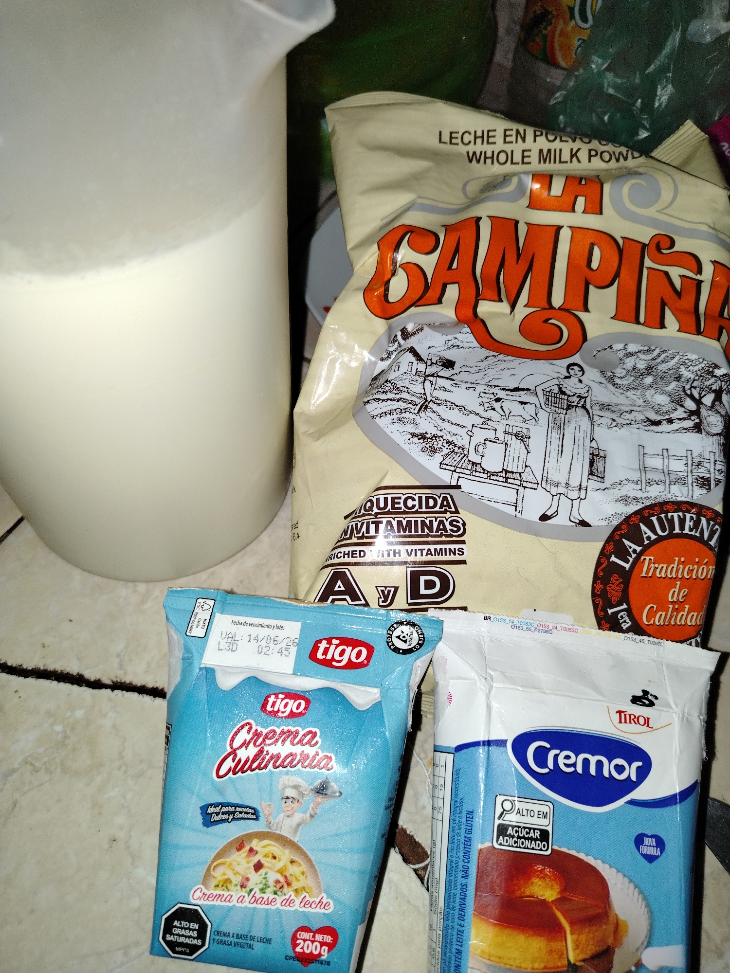 This close-up indoor shot displays various food ingredients on a light-colored tiled surface, suggesting a kitchen setting in Tinaquillo, Venezuela.
To the left, a large white plastic pitcher contains a quantity of white liquid, likely milk, with some frothing visible on the surface.
Dominating the center is a large, beige-colored bag of "LA CAMPINA" brand powdered milk. The packaging features the text "LECHE EN POLVO" (Powdered Milk) and "WHOLE MILK POWDER" at the top. The prominent orange brand name "LA CAMPINA" overlays an illustration of a rural scene with a woman, a man, and farm animals. Further text indicates it is "ENRIQUECIDA CON VITAMINAS A y D" (Enriched with Vitamins A and D) and proclaims "LA AUTÉNTICA Tradición de Calidad" (The Authentic Tradition of Quality).
At the bottom left, a blue carton of "tigo Crema Culinaria" (Culinary Cream) is visible. It features a cartoon chef and a dish of pasta. Text on the package states "Ideal para recetas Dulces y Saladas" (Ideal for sweet and savory recipes), "Crema a base de leche y grasa vegetal" (Cream made from milk and vegetable fat), and highlights "ALTO EN GRASAS SATURADAS" (High in Saturated Fats). The net content is "CONT. NETO: 200g". A visible expiration date reads "VAL: 14/06/26 L3D 02:45".
To the bottom right, partially obscured, is a blue packet of "TIROL Cremor" dessert mix. The packaging displays an image of flan or pudding and is marked "ALTO EM AÇÚCAR ADICIONADO" (High in Added Sugar) and "NOVA FORMULA" (New Formula). The side of the packet mentions "CONTÉM LEITE E DERIVADOS. NÃO CONTÉM GLUTEN." (Contains milk and derivatives. Does not contain gluten.).
The arrangement of ingredients suggests preparation for cooking or baking. The lighting is even, and no people or specific time of day or emotional cues are discernible.