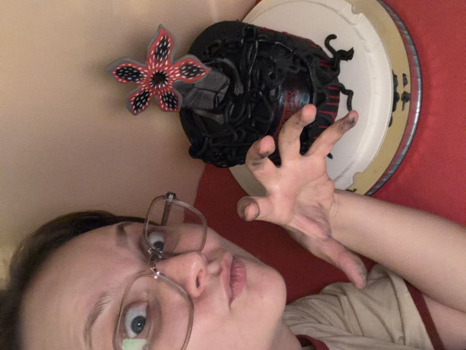A person wearing glasses poses next to a black cake with a demogorgon head and flower, reminiscent of "Stranger Things". Their hand is outstretched with fingers slightly curled and fingertips smudged with black. The cake sits on a white and gold base against a red background. The person looks directly at the camera with a slight tilt of the head. They are wearing a beige shirt with a red collar.