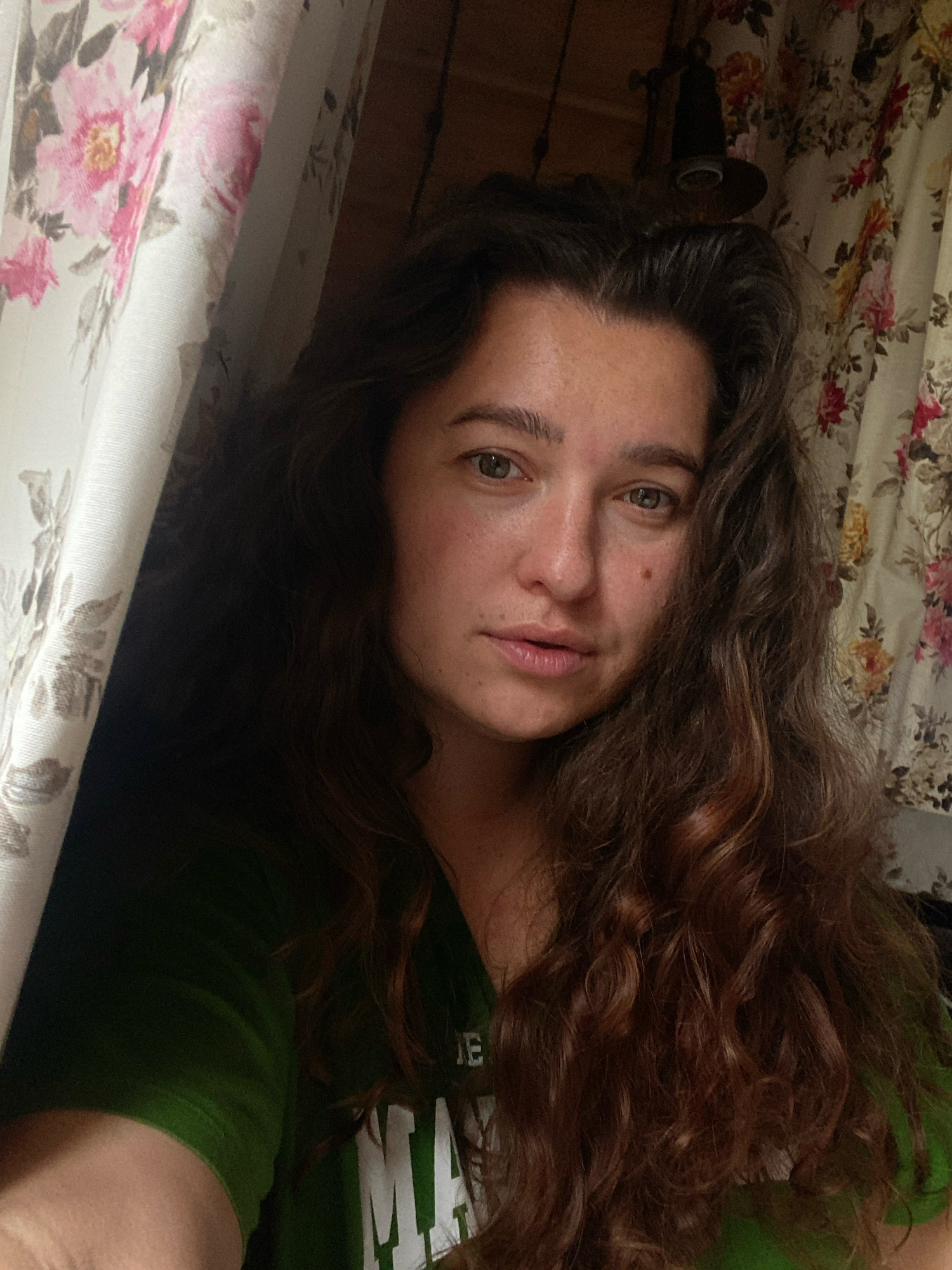 A self-portrait of a woman with long, wavy brown hair. She is wearing a green t-shirt with white lettering that is partially visible. The woman is looking directly at the camera with a neutral expression. She is positioned in front of a curtain with a floral pattern, featuring pink and yellow flowers. To the left of the woman, a portion of another curtain with a similar floral pattern is visible. The background appears to be a room with wooden walls. The image does not provide any information or visual cues that can be used to determine the location in the city of Perm, Russia.
