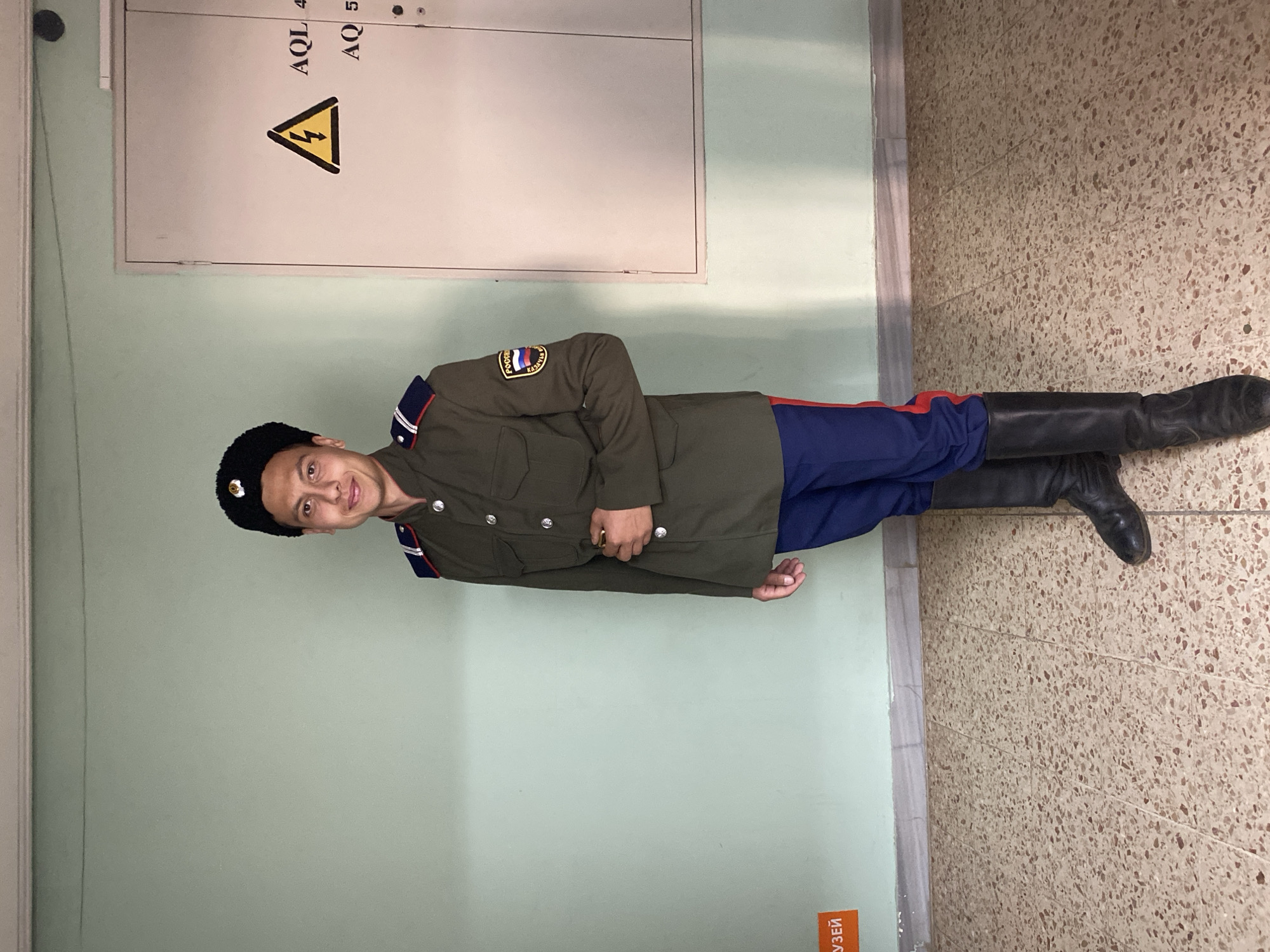 A man in a Cossack uniform stands in front of a wall, likely inside a building or museum. He is wearing a black Kubanka hat, a dark green tunic with a Russian flag patch, blue pants with a red stripe, and black leather boots. The wall behind him has a light green color on the bottom and a gray panel at the top, containing an electricity warning symbol and the letters "AQL" and "AQ." The floor next to the wall is tiled with beige and brown speckled tiles. Part of an orange sign with Russian writing can be seen in the lower left corner, possibly a museum or exhibit label. The location is Markovskii, Russia.