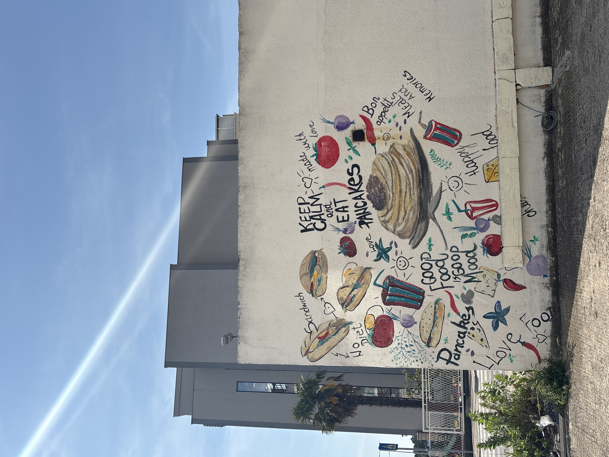 An outdoor shot captures a vibrant, food-themed mural painted on a white building wall in Bar, Montenegro, under a clear, bright blue sky with a visible sunbeam. The mural features a large stack of pancakes as a central element, surrounded by illustrations of various other food items such as sandwiches or subs, vegetables like tomatoes, onions, and chili peppers, cheese, and colorful beverages in striped cups.
Interspersed throughout the mural are numerous hand-drawn texts and phrases. Key phrases include "KEEP CALM and EAT PANCAKES made with love," "Bon appetit," "MEALS And Memories," "Happy food," "GOOD Food GOOD Mood," "Pancakes," "Love Food," "Sandwich," and "No Diet." Smaller hearts and star shapes also decorate the artwork.
To the left of the mural, a modern gray building rises, with a palm tree visible at its base. The ground beside the painted wall is a light-colored, possibly concrete, path, and some small green bushes grow near the bottom right. A security camera is mounted on the white wall near the gray building, and a coiled wire is visible further down the wall's edge. The scene is brightly lit, indicating daytime and sunny weather.